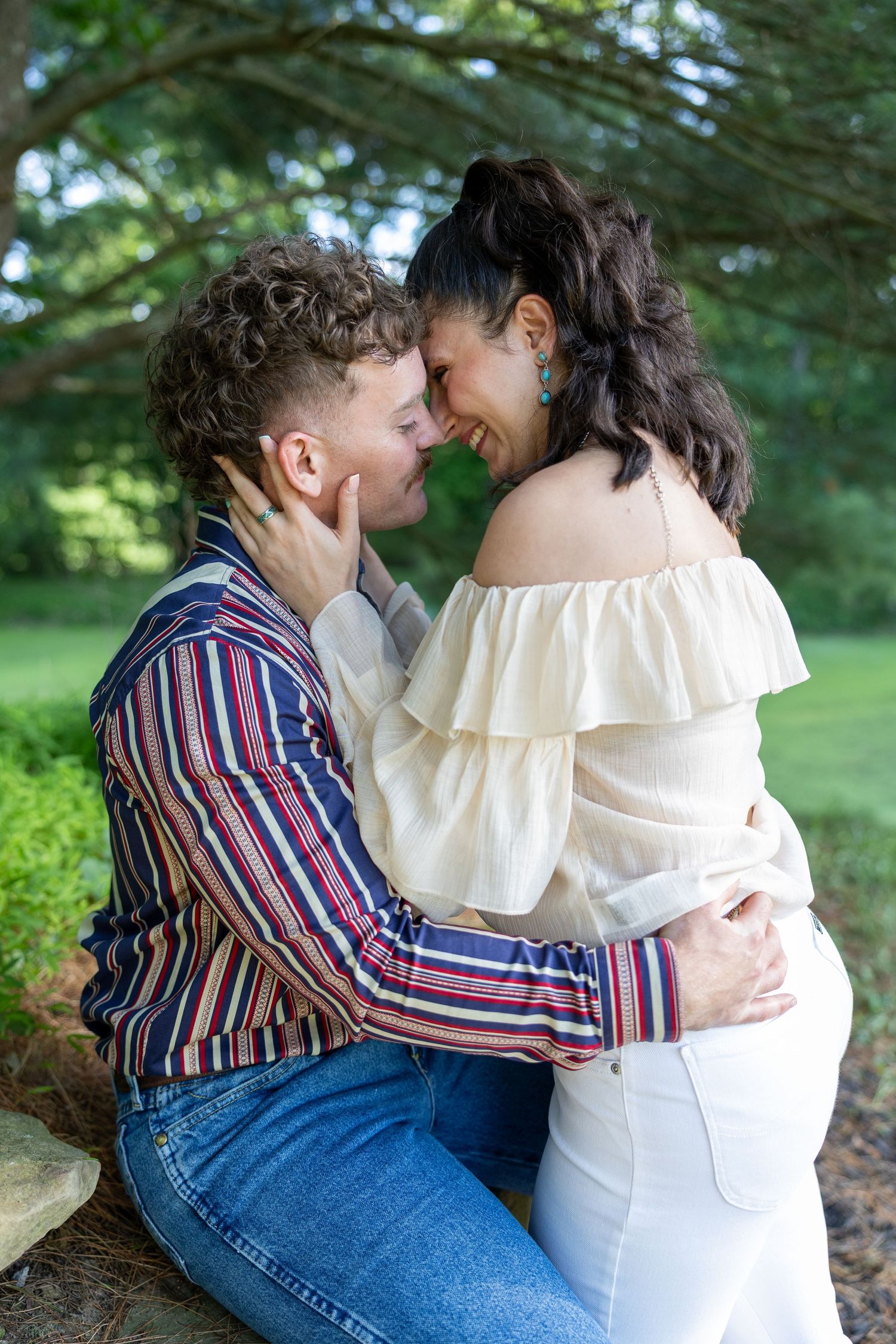Couple sharing tender nose-to-nose moment outdoors, woman in off-shoulder top and white pants.