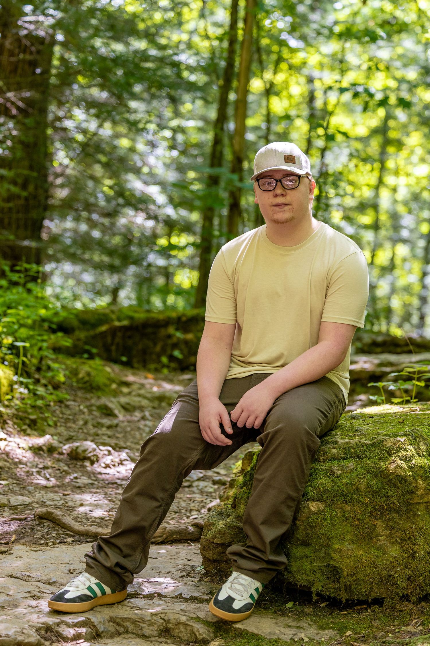 Young man in yellow t-shirt, cap, and glasses sitting on mossy rock in lush green forest setting.
