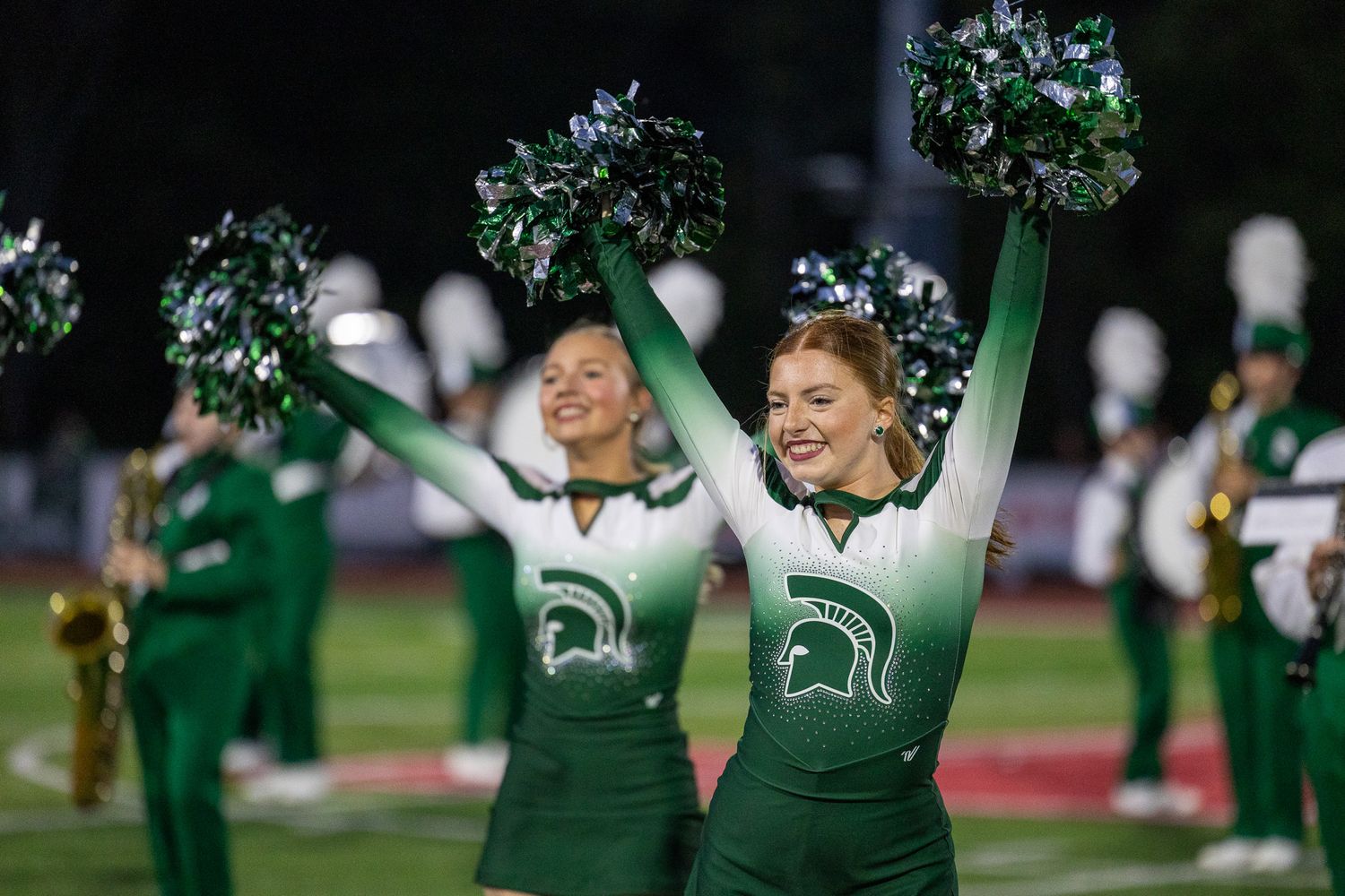 Two cheerleaders in green and white uniforms wave pom-poms on the field during a night football game.