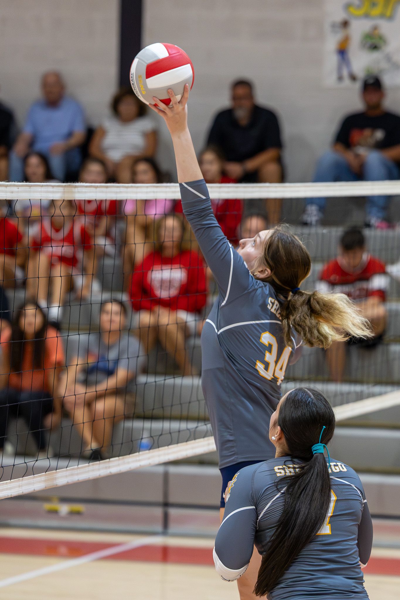 Player #34 reaches up to tip a volleyball over the net during a competitive indoor volleyball match with fans watching.