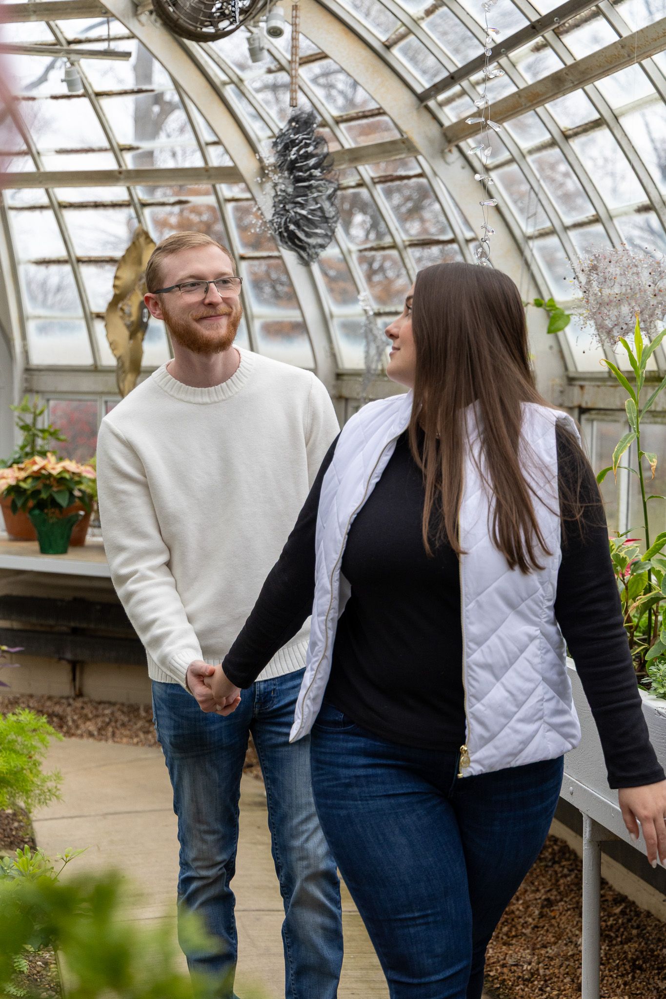 Couple holding hands and smiling at each other inside a sunlit greenhouse with lush green plants.