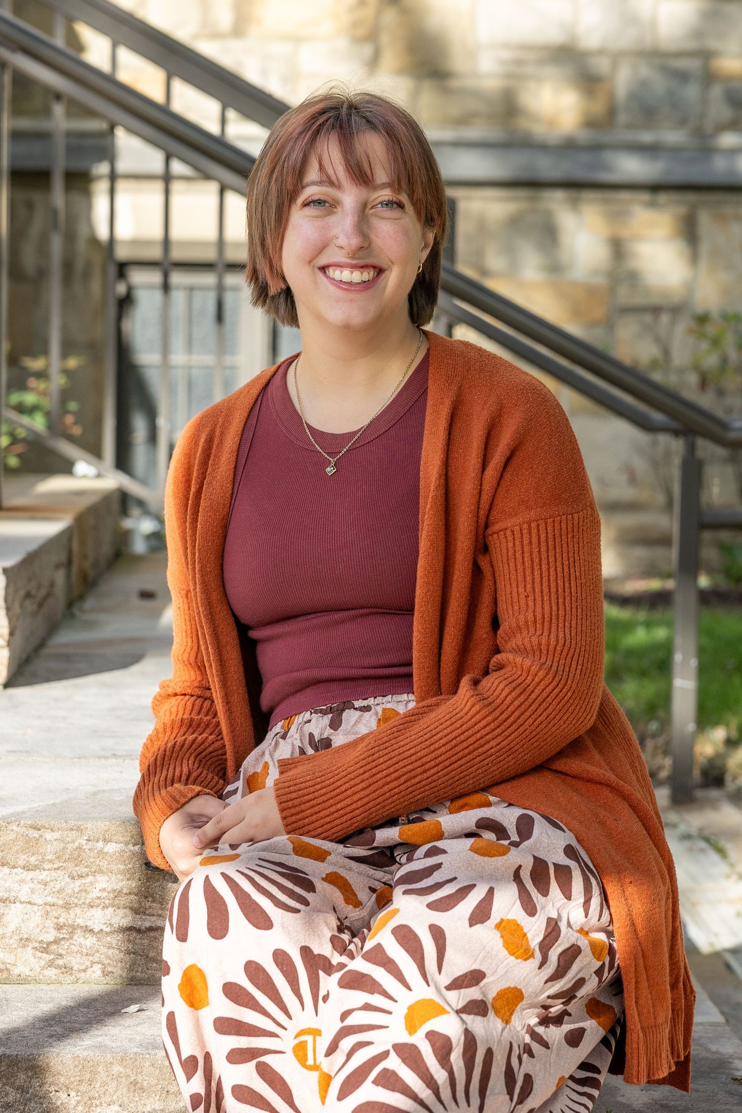 Smiling young woman in orange cardigan and floral skirt sitting on stone steps outdoors.