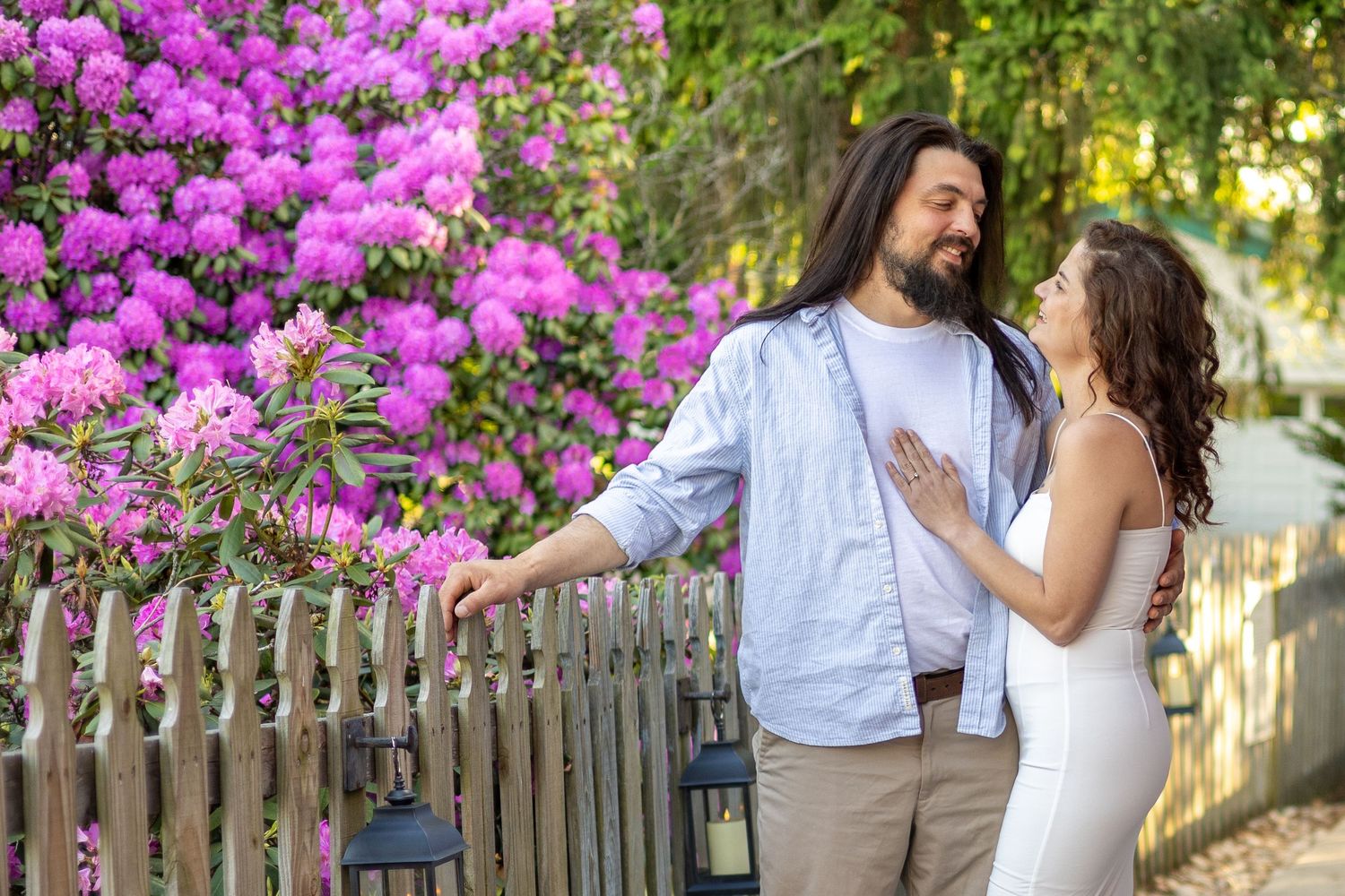 Couple smiling near purple flowering bushes by a wooden fence.