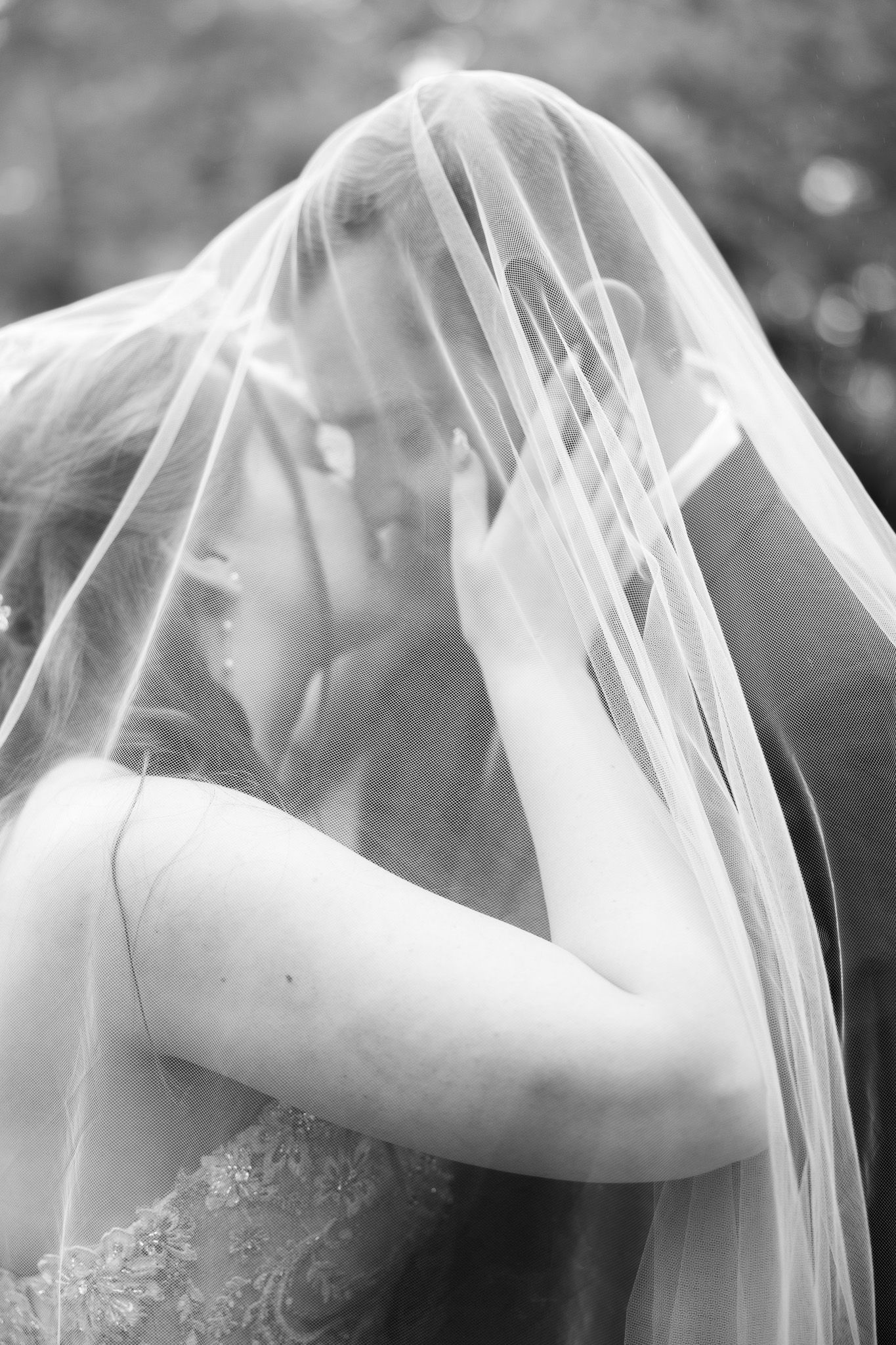 Black and white romantic photo of bride and groom kissing under flowing wedding veil outdoors.