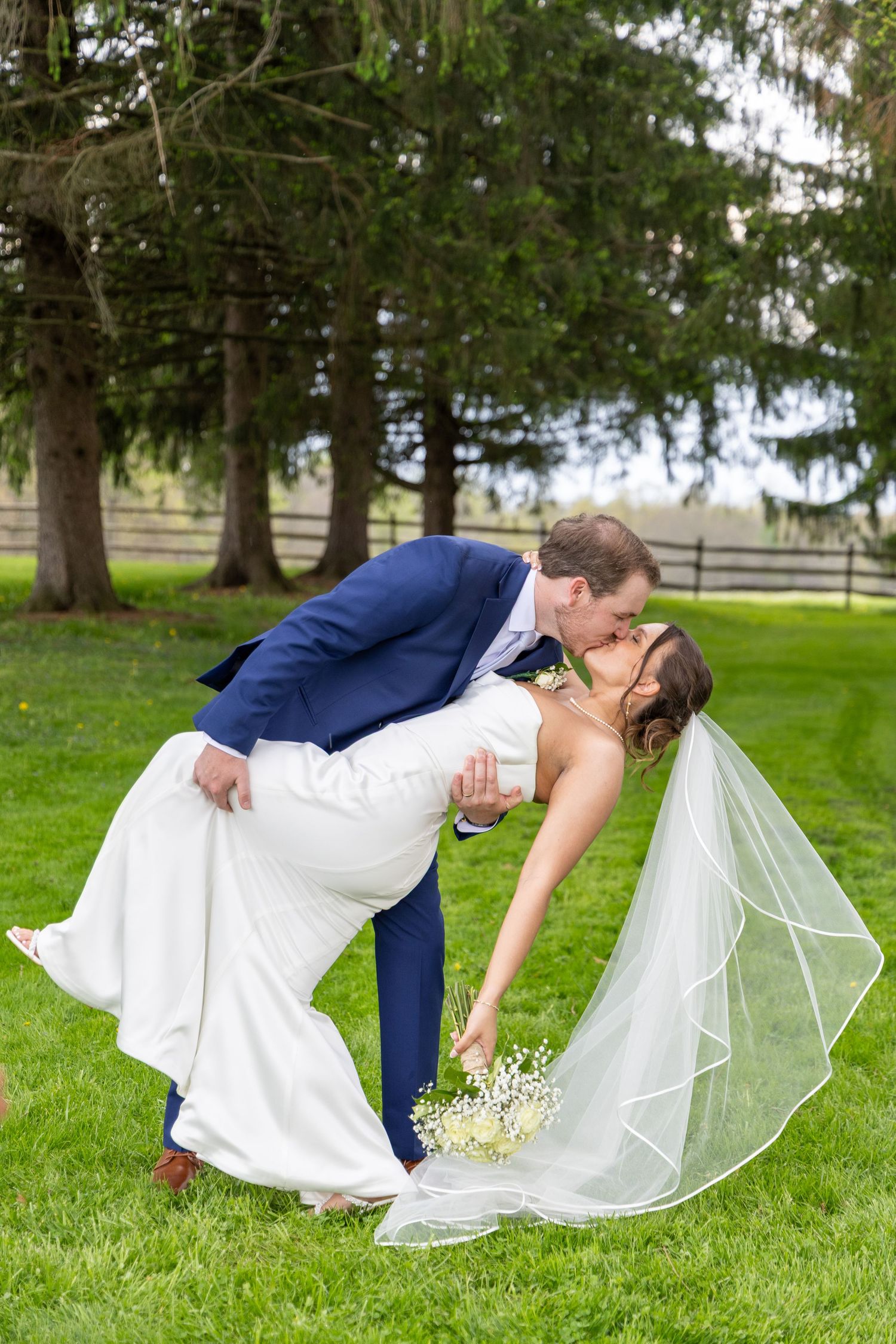 Groom in blue suit dips bride in white gown and veil for a romantic kiss on lush green lawn.