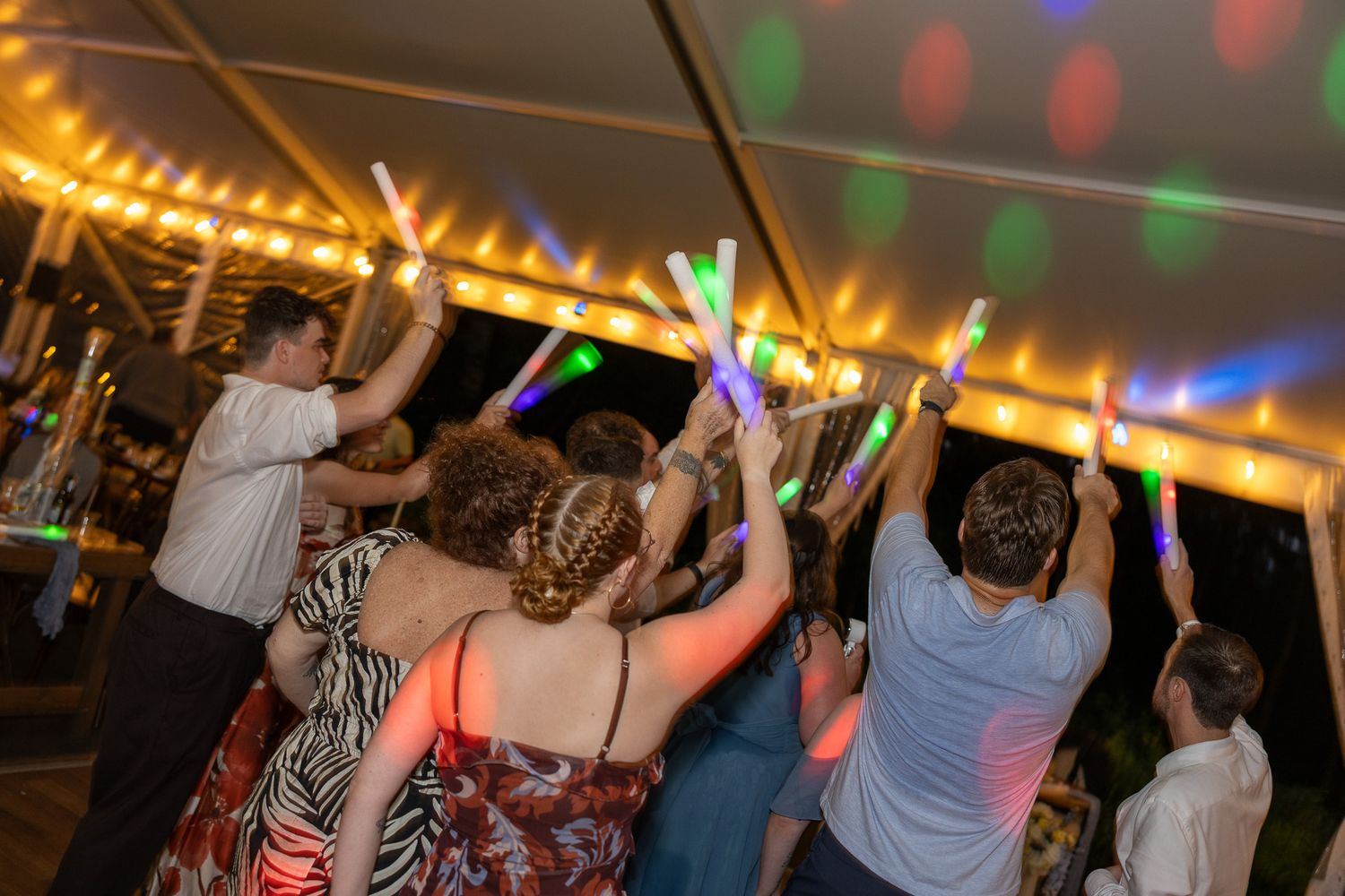 People dancing at a party under string lights, holding colorful glowing foam sticks in a festive outdoor tent.