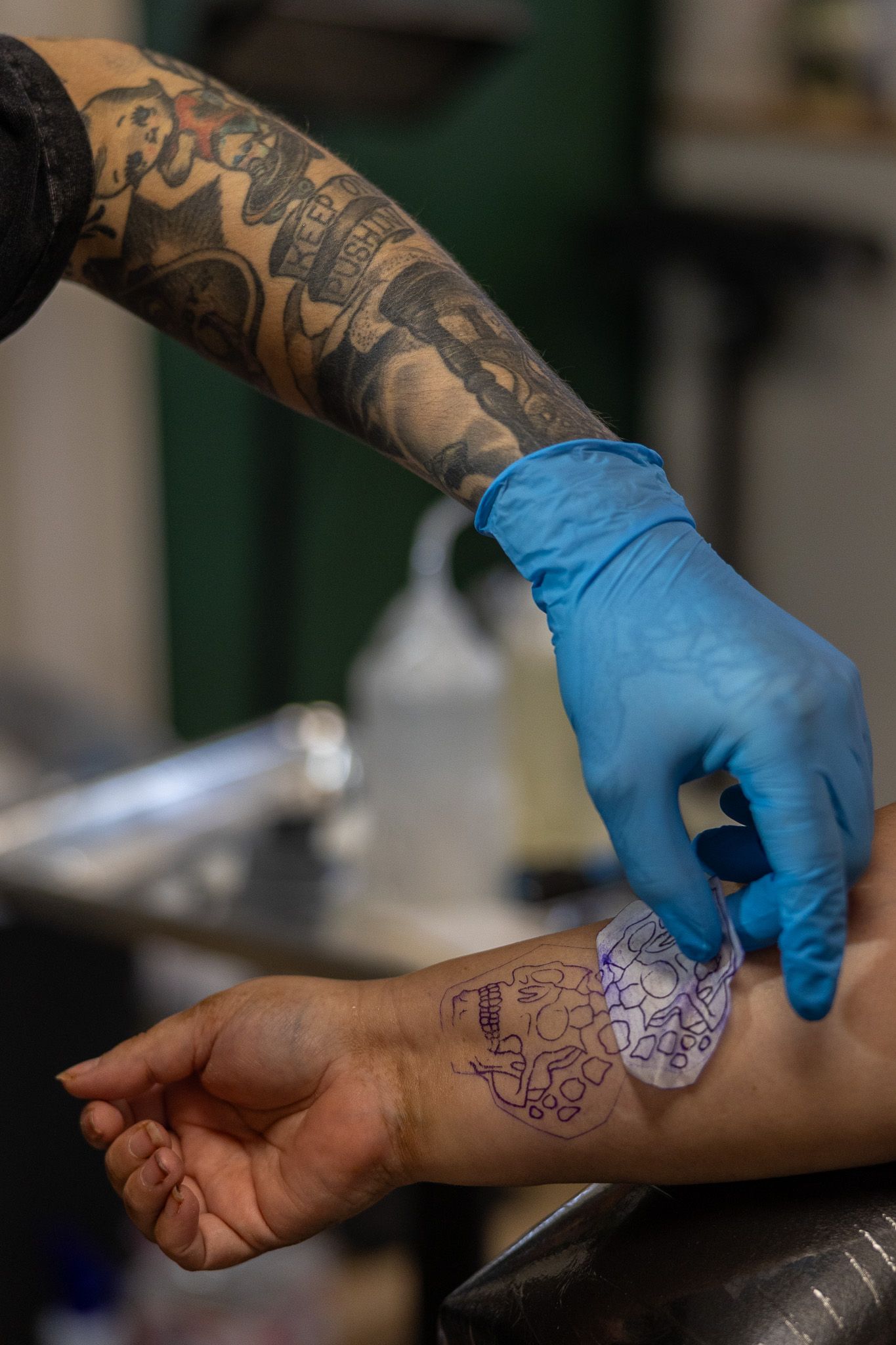 Tattooed artist wearing blue gloves applies a stencil transfer onto a clients wrist in a tattoo studio.