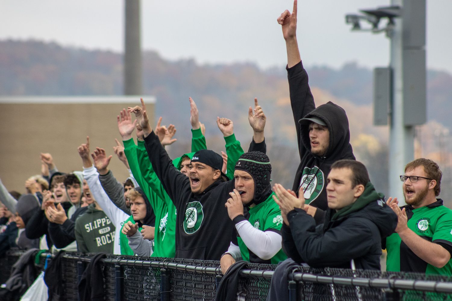 Slippery Rock University fans in green cheer enthusiastically at an outdoor college football game in autumn.