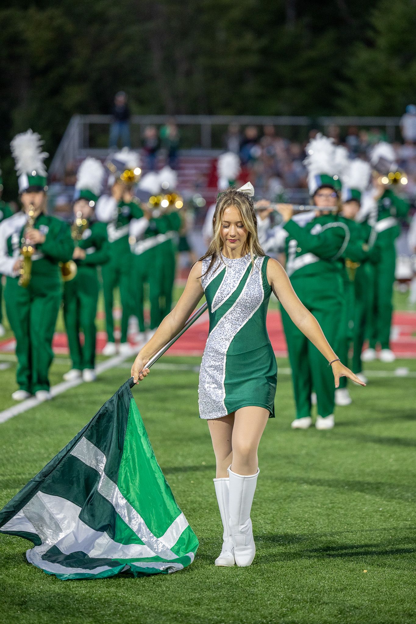 A female color guard performer in a green and silver uniform twirls a flag on a football field with a marching band behind her.