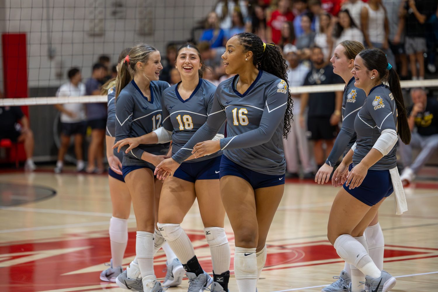 College volleyball players in blue uniforms celebrate together on the court during a competitive indoor match.