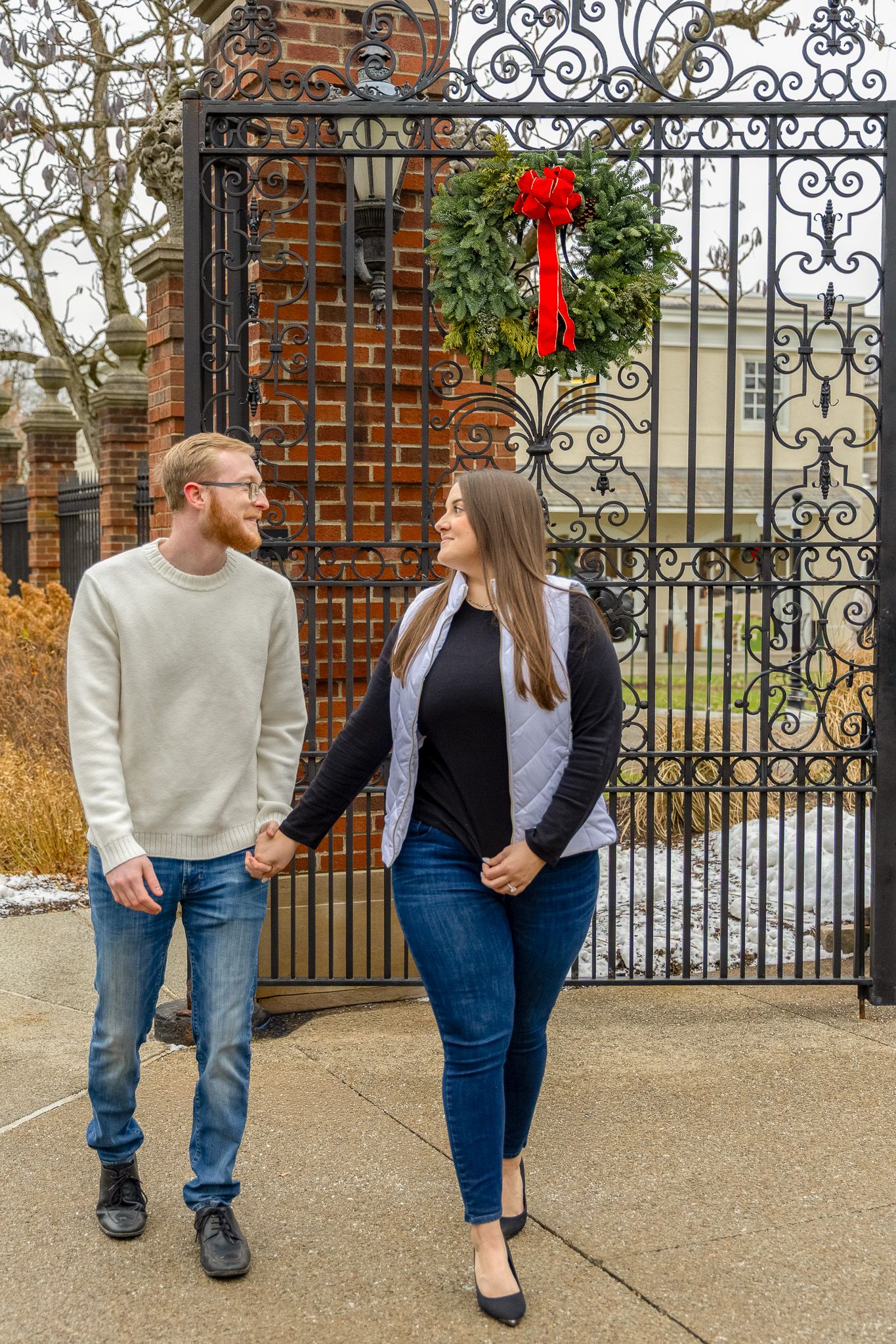 A couple holds hands and smiles at each other before an ornate iron gate decorated with a Christmas wreath.