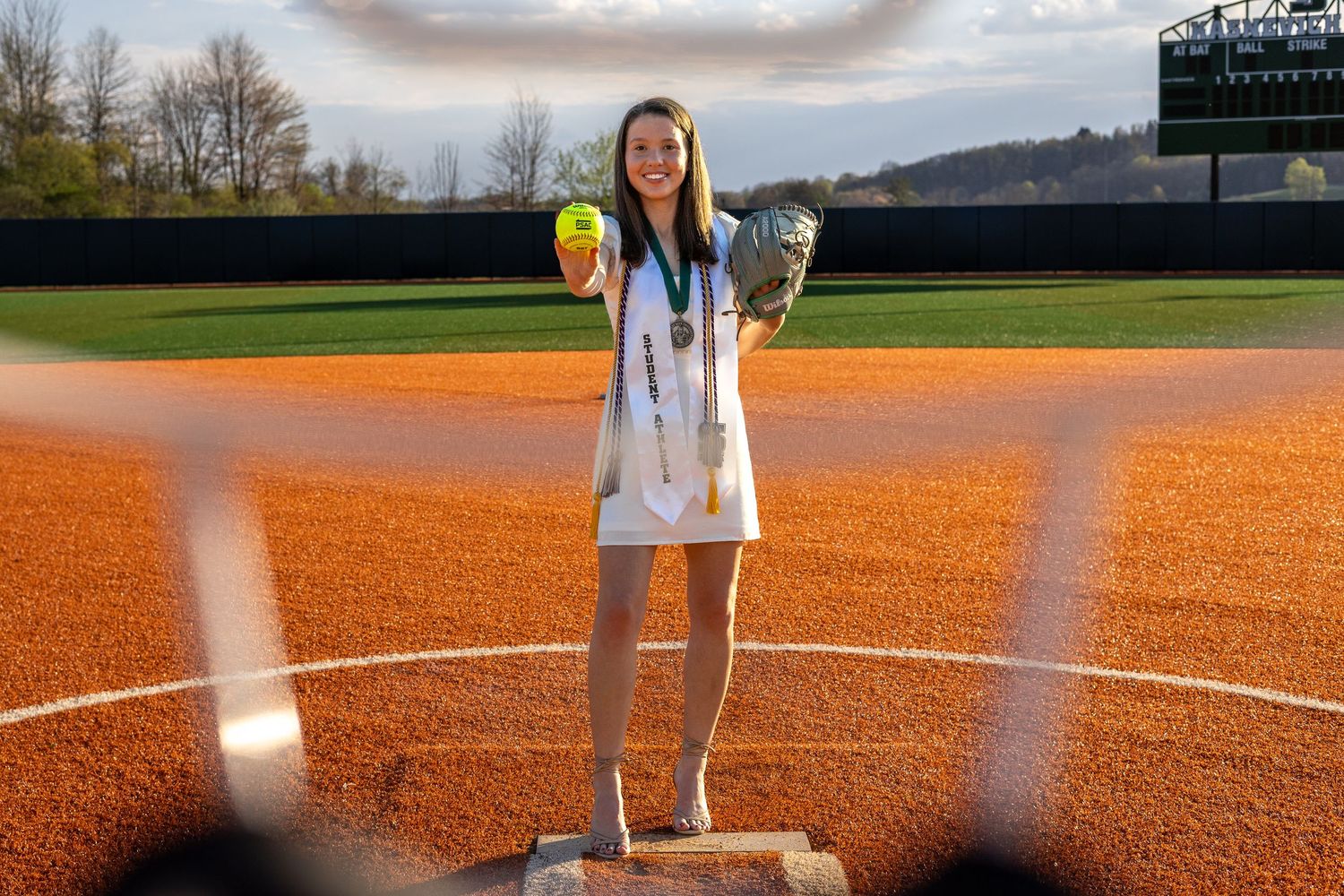 Female softball player in graduation attire holds ball and glove while standing on pitcher's mound.