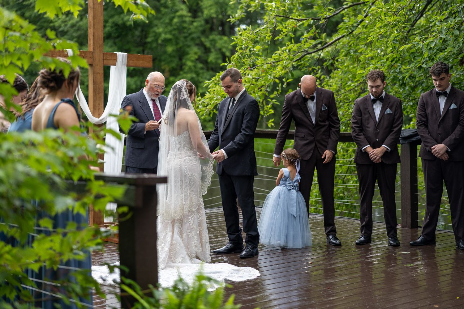 Bride and groom hold hands during outdoor rainy wedding ceremony with flower girl and groomsmen nearby.