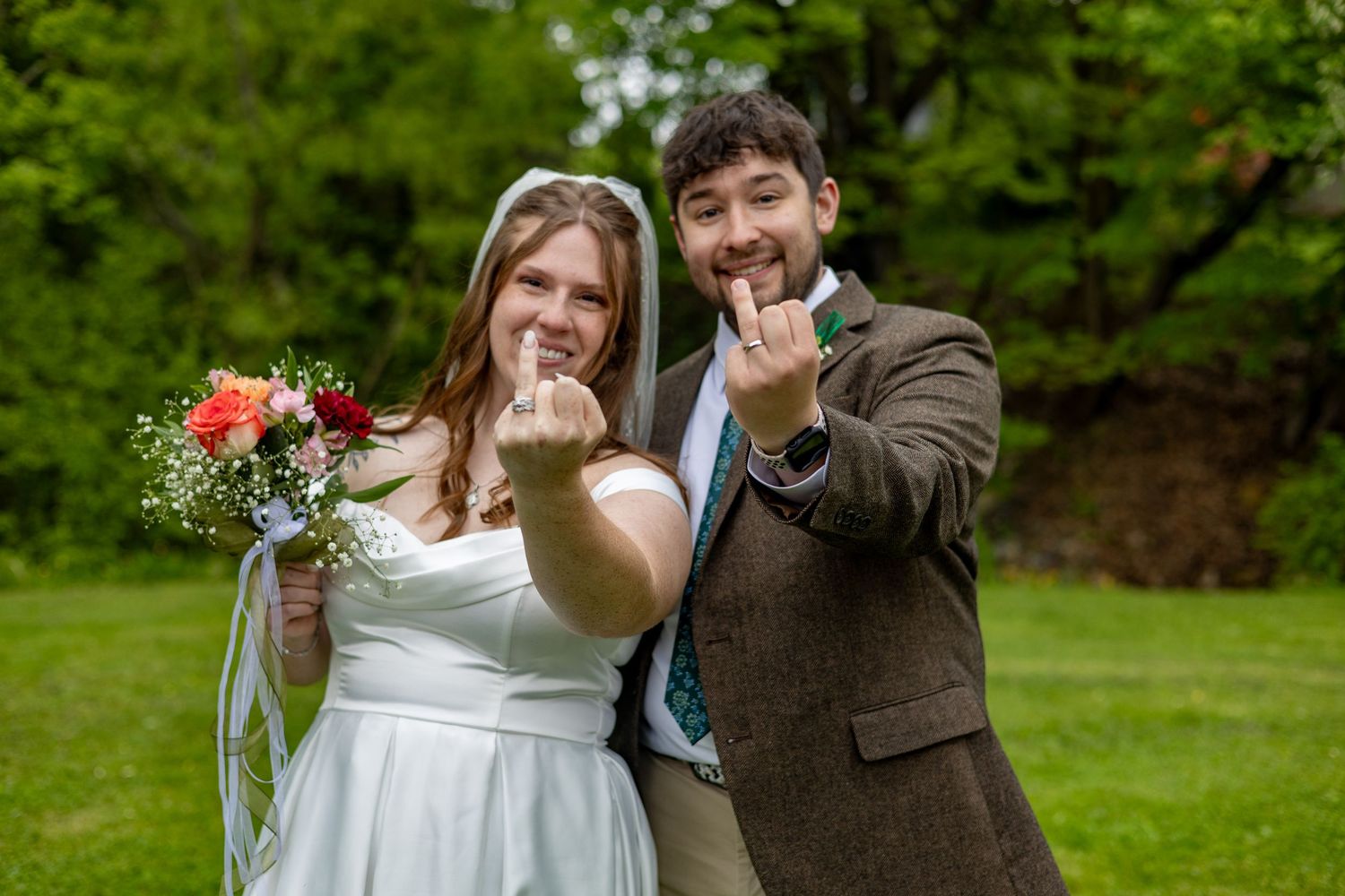 Newlyweds smiling and showing off wedding rings, bride holding colorful bouquet in green outdoor setting.