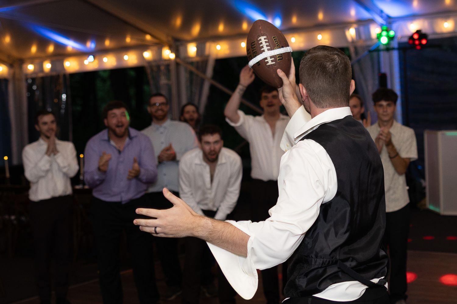 Groom tossing a football to groomsmen during a wedding reception dance floor celebration under string lights.