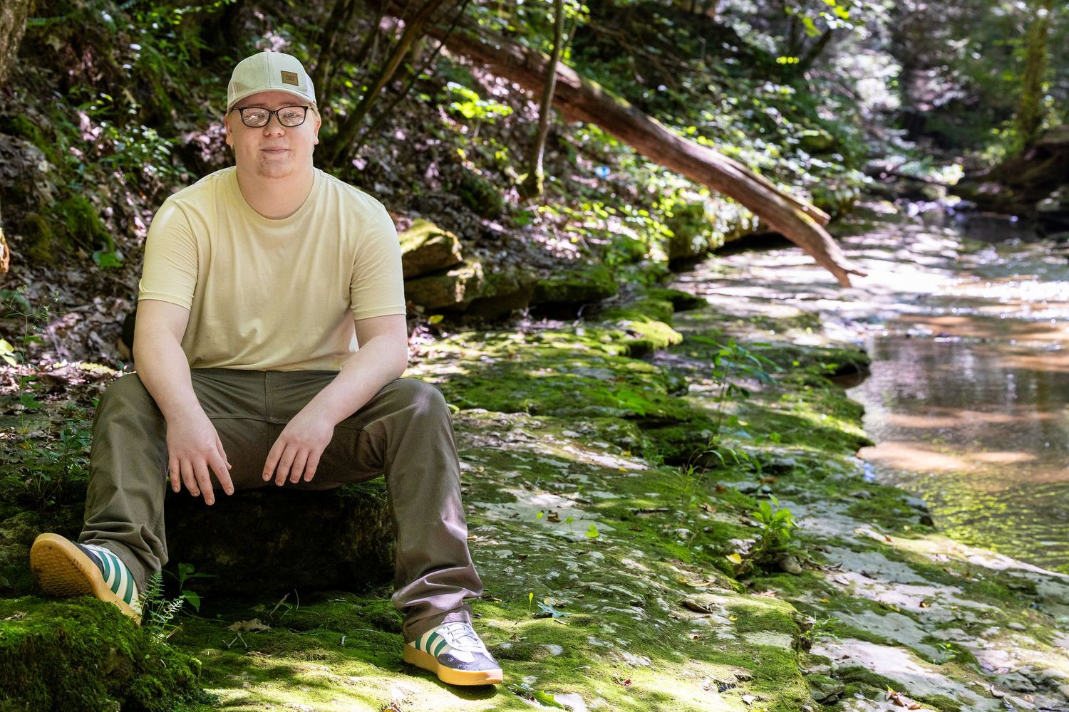 Person in green hat and olive shirt sits on mossy rock beside a serene forest creek surrounded by lush greenery.