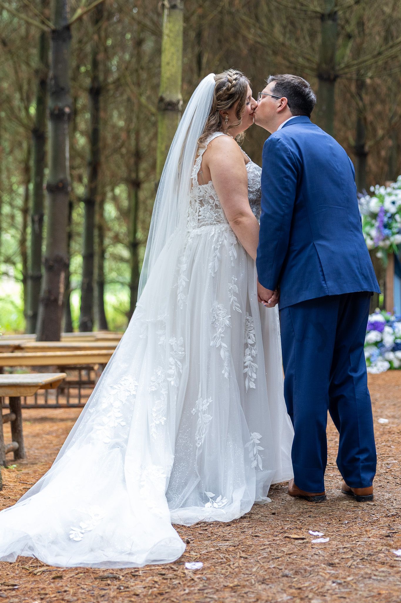 Bride and groom share first kiss at outdoor forest wedding ceremony, bride in lace gown with veil, groom in blue suit.