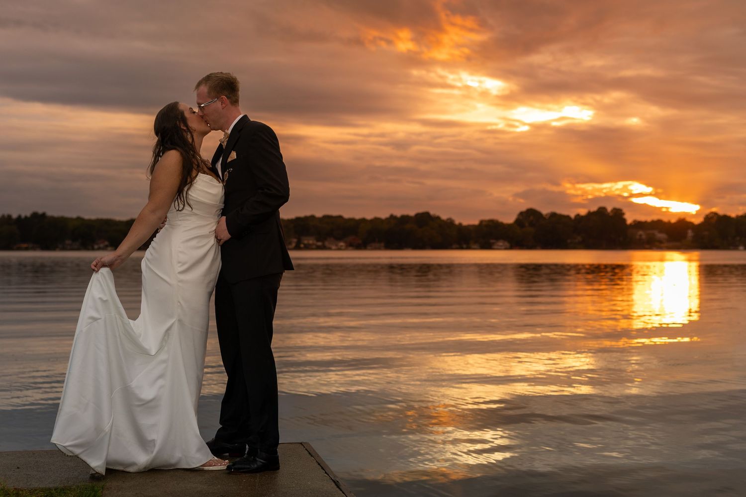 Newlyweds share a romantic kiss by a lakeside at sunset, with vibrant orange skies reflecting on the water.