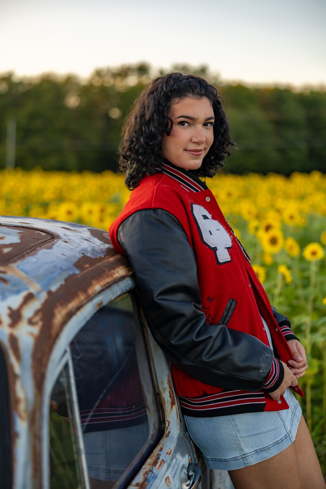 Teen girl in red varsity jacket leans on rustic car in front of vibrant sunflower field at sunset.