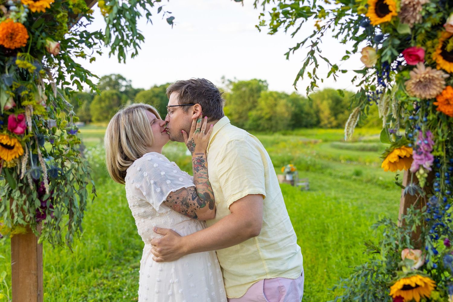 Couple sharing a kiss under a floral arch with sunflowers at an outdoor wedding ceremony in a field.