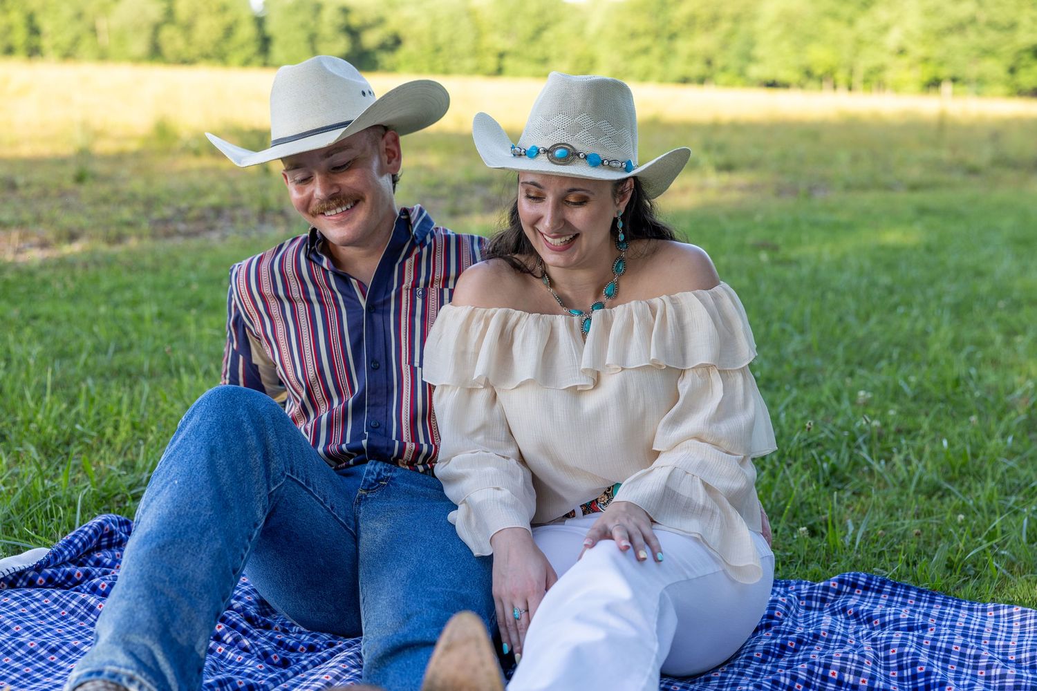 Couple in cowboy hats sitting on a blue checkered blanket in a green field, smiling and relaxed.
