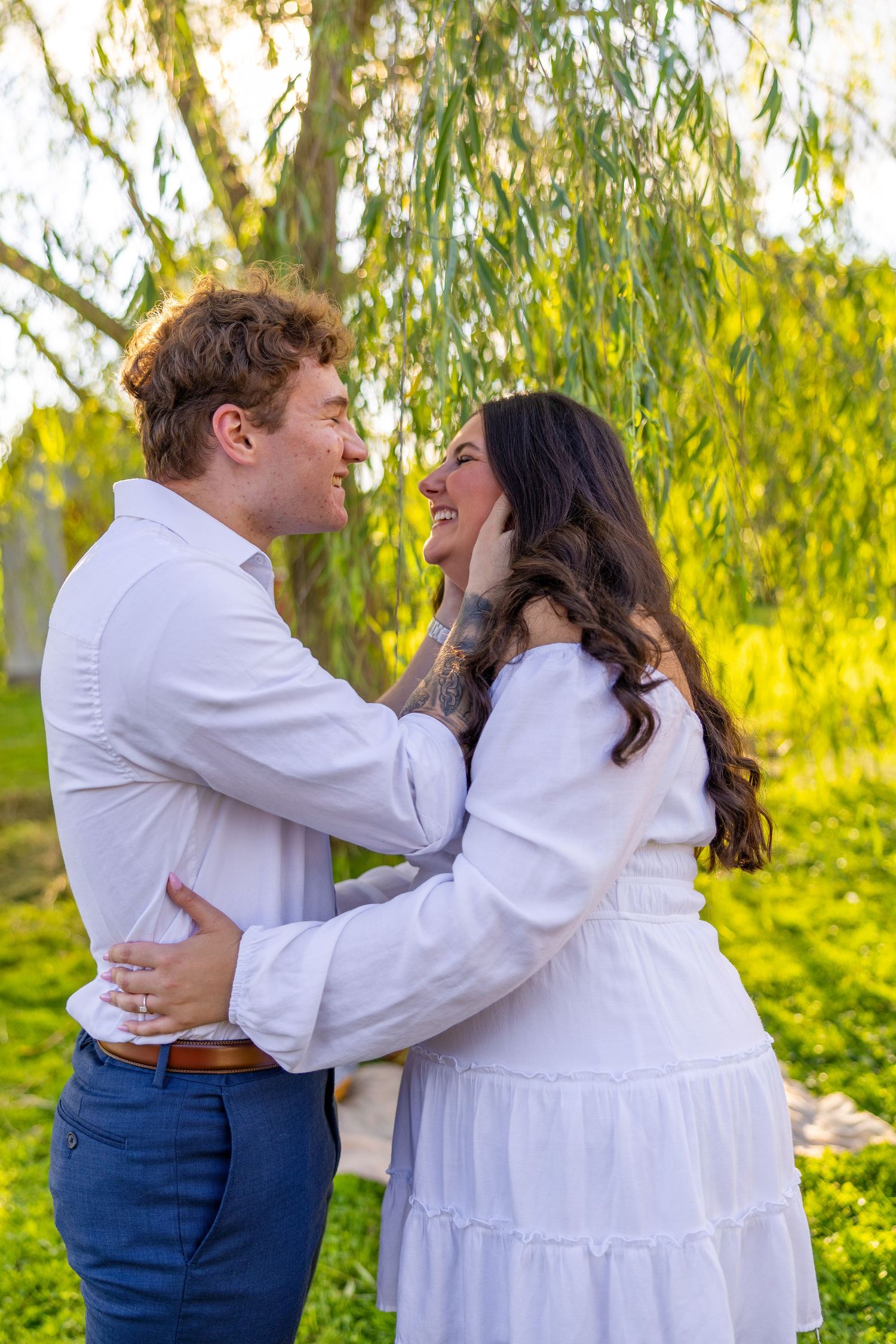 Romantic couple embraces and smiles at each other under weeping willow tree during engagement photos.