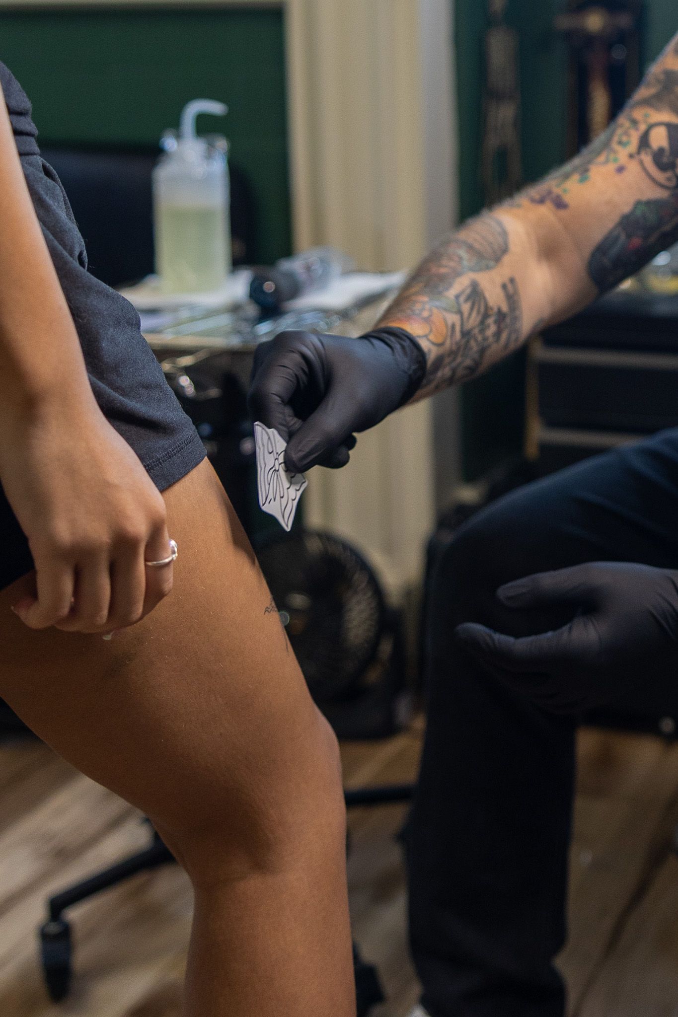 Tattooed artist in black gloves placing a stencil on a client's thigh in a tattoo studio.