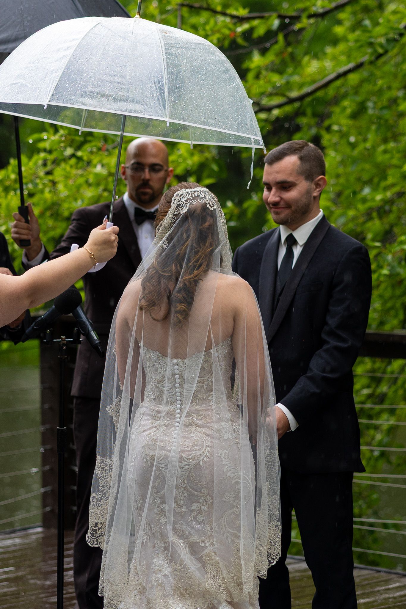 Groom smiles at veiled bride under clear umbrella during intimate rainy outdoor wedding ceremony.