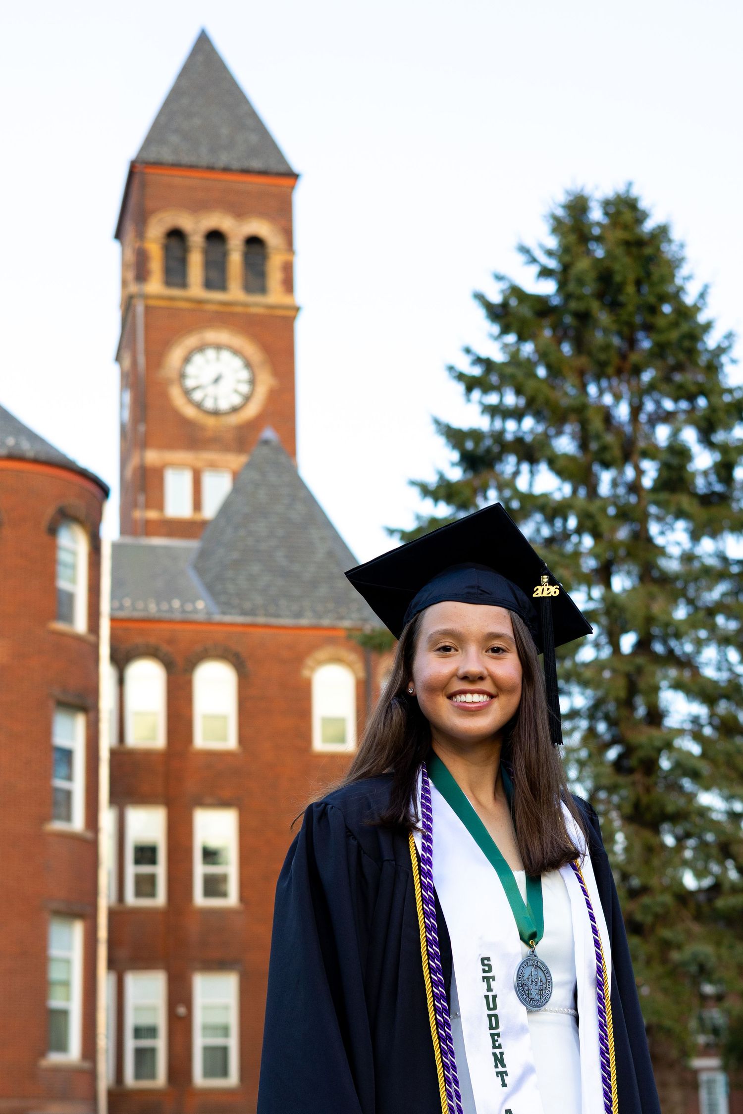 Female graduate in cap and gown with honor medal stands before Slippery Rock University clock tower.