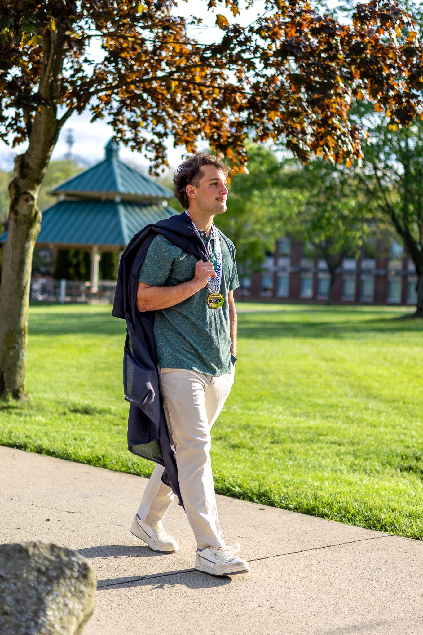Male graduate carries gown over shoulder while walking through sunny Slippery Rock University campus.