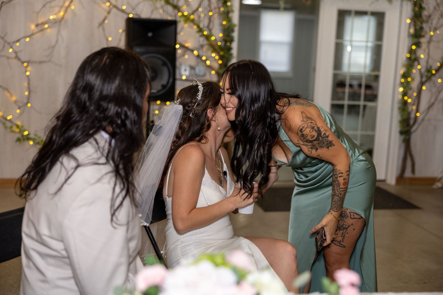 Bride in white gown and veil shares a tender moment with tattooed bridesmaid in sage green dress at elegant wedding reception.