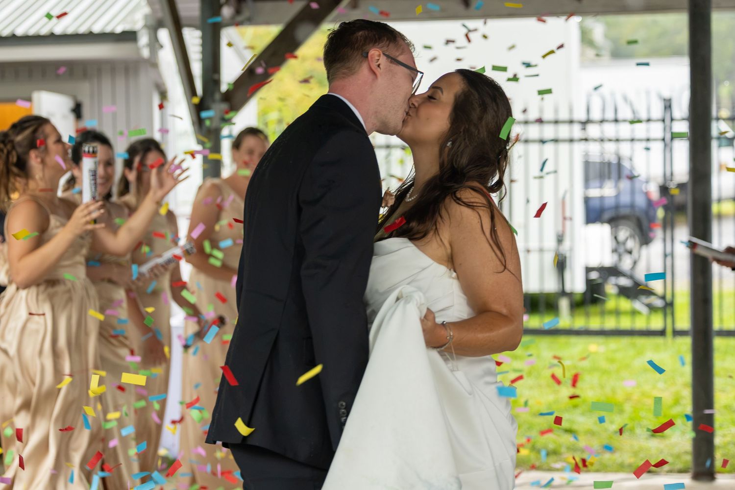 Newlywed couple sharing a kiss surrounded by colorful confetti as bridesmaids celebrate at outdoor wedding ceremony.