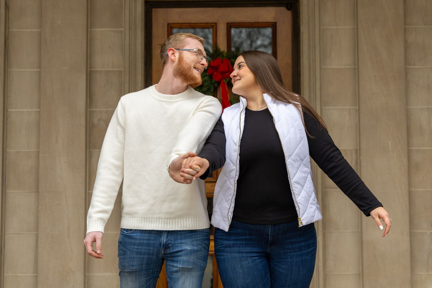 A smiling couple holds hands in front of a decorated door, wearing casual winter outfits with a holiday wreath backdrop.