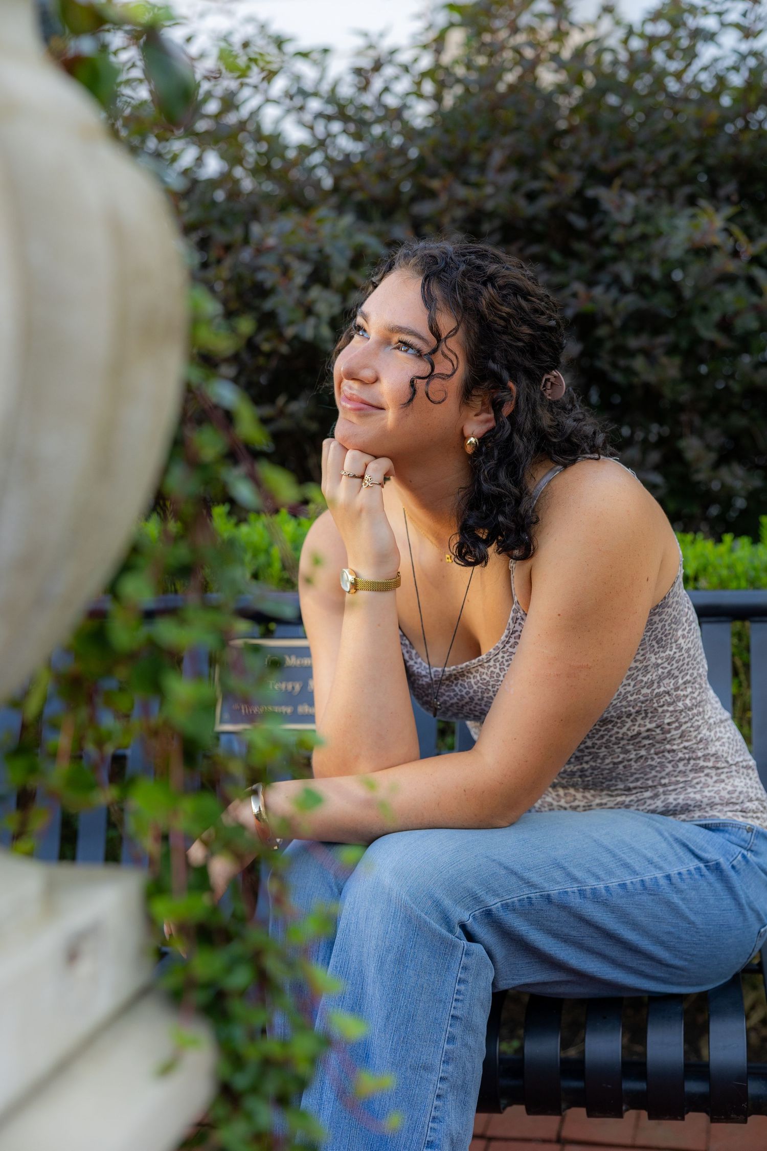 Young woman with curly hair sits on a bench in a garden, resting her chin on her hand, smiling thoughtfully.