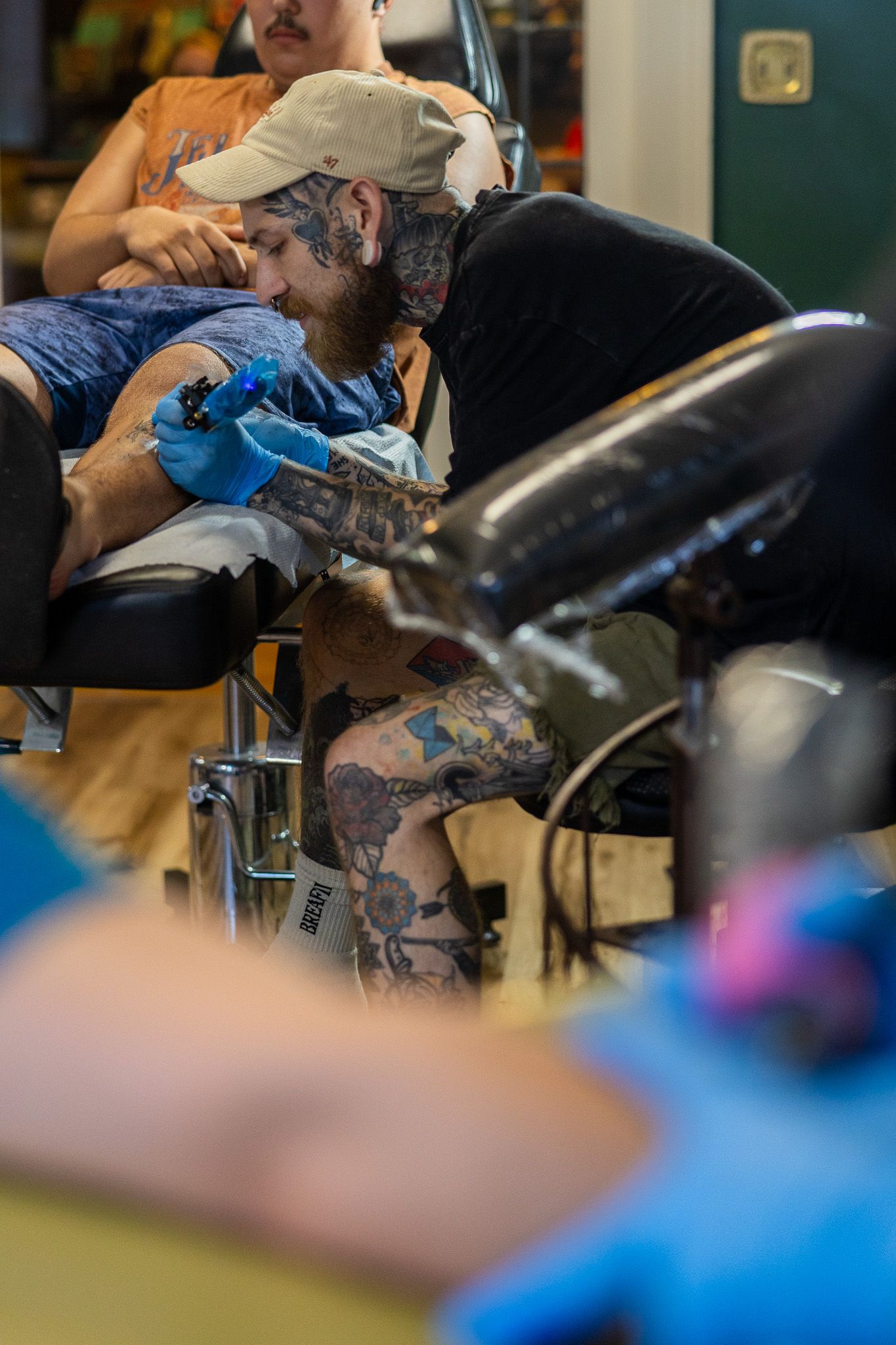Tattooed artist wearing gloves carefully tattoos a client's arm in a busy professional tattoo parlor.