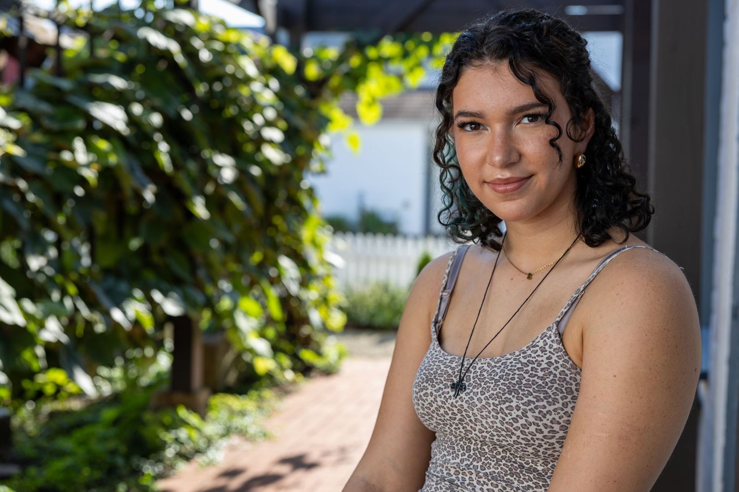 Young woman with curly hair wearing a floral top and necklace, posing outdoors in a sunny garden setting.