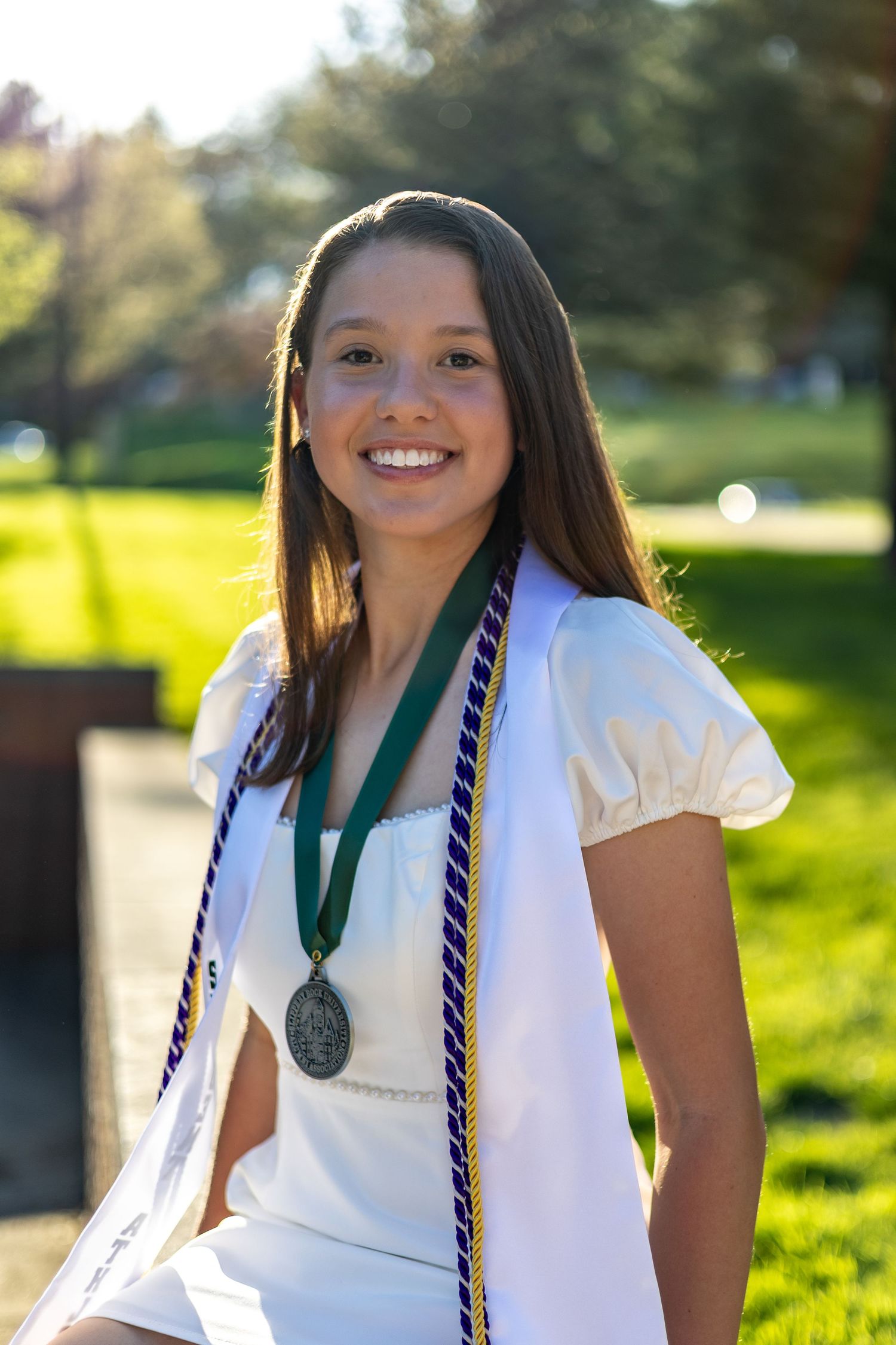 Smiling young woman in white dress wearing graduation medal and purple gold honor cords seated outdoors.