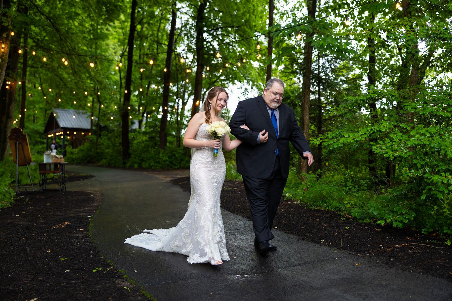Bride in lace gown walks down rainy forest path with escort under string lights before ceremony.