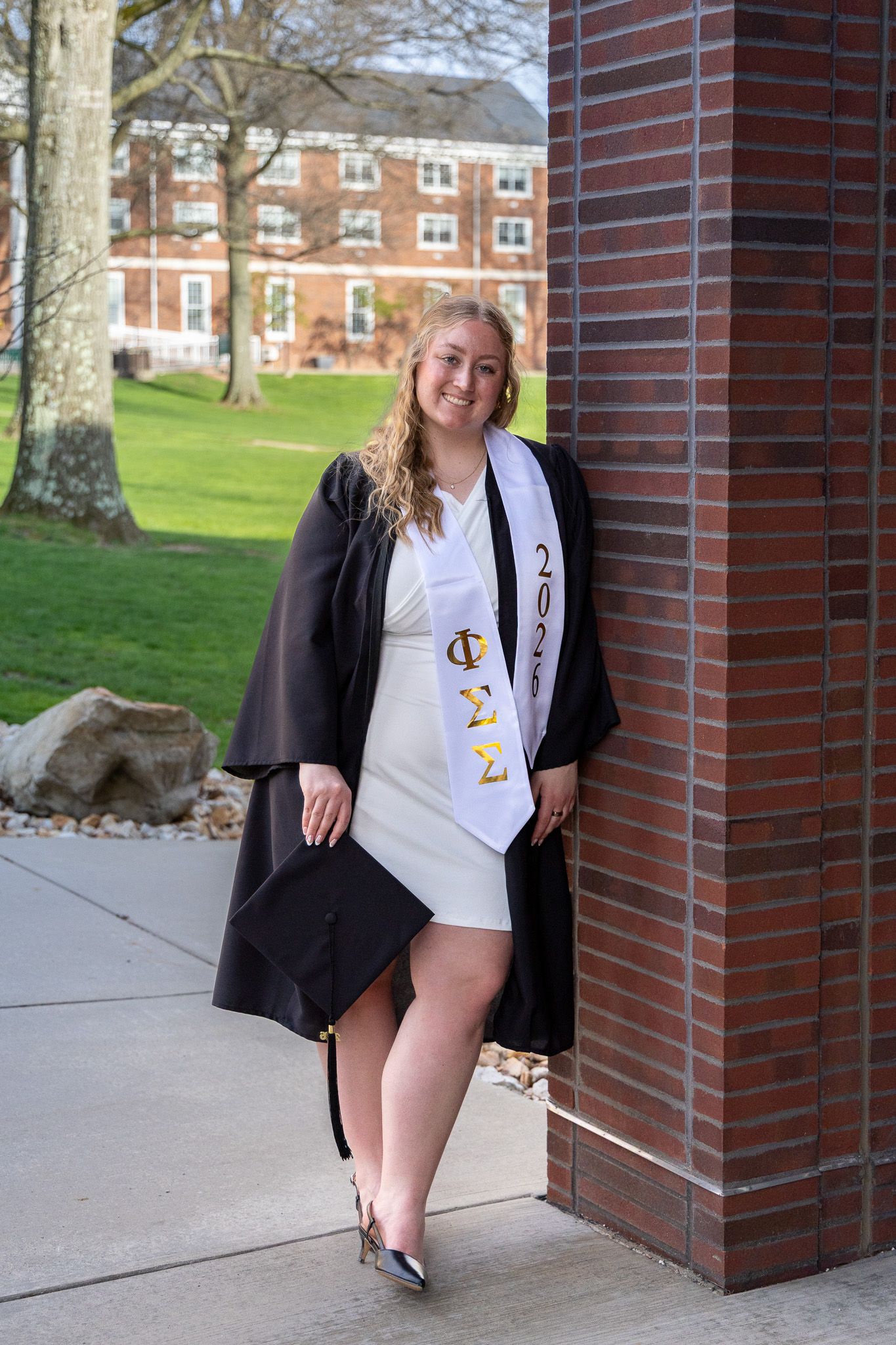 Female graduate in cap and gown with Chi Sigma sorority stole poses by brick column.