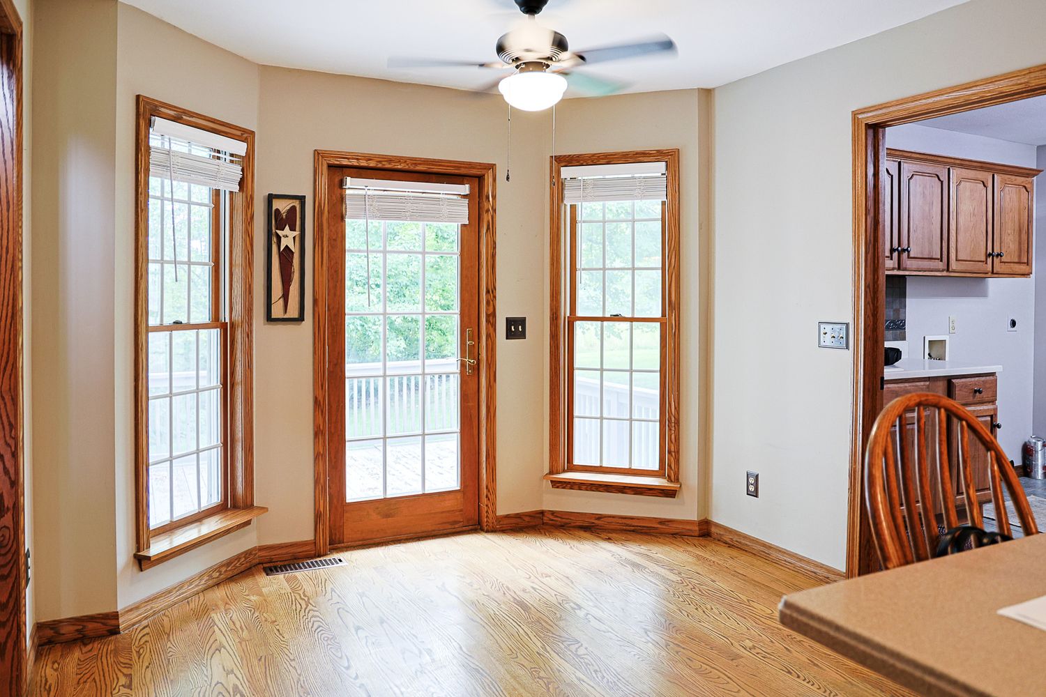 Bright dining area with hardwood floors, bay windows, ceiling fan, and wood trim leading to kitchen.