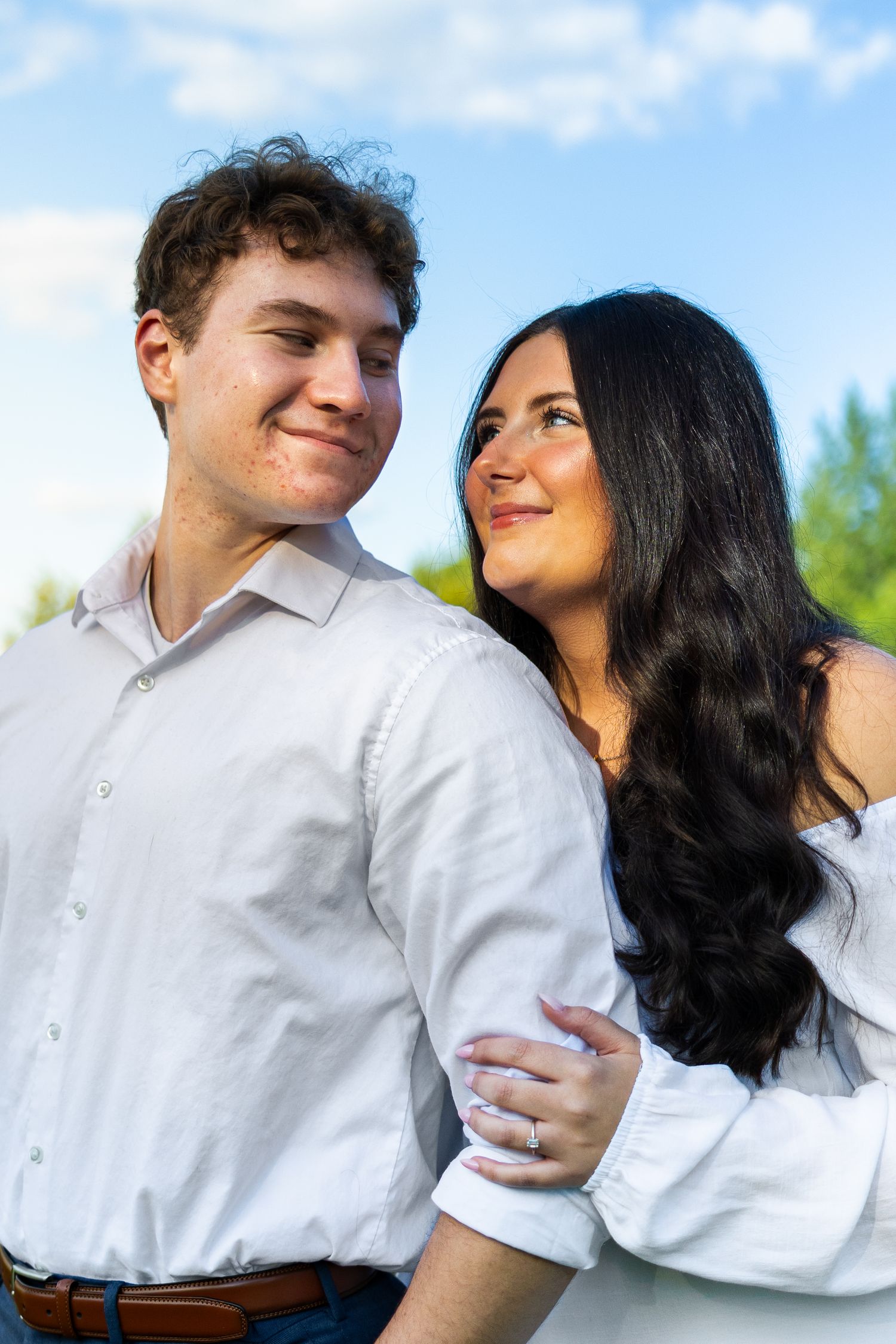 A young couple in white shirts embrace outdoors, smiling under a blue sky with green trees behind them.