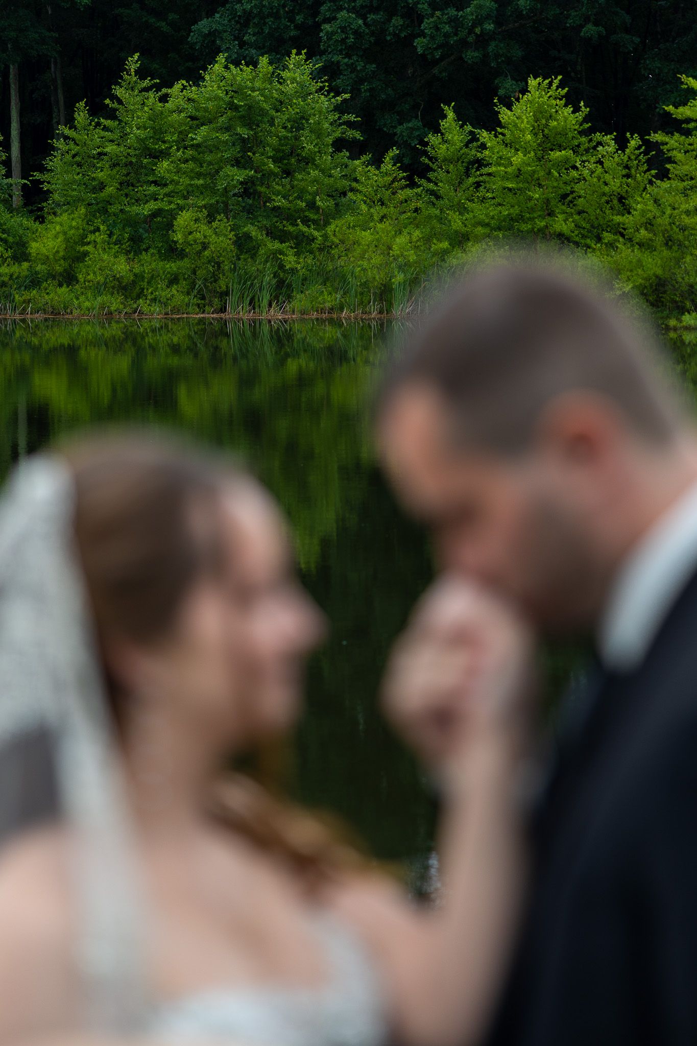 Artistic blurred foreground of bride and groom near reflective lake surrounded by lush green trees.