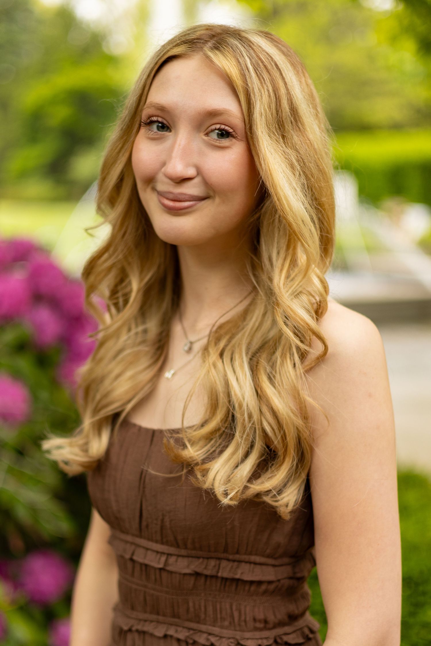 Smiling teenage girl with long blonde wavy hair posing outdoors near pink flowers.