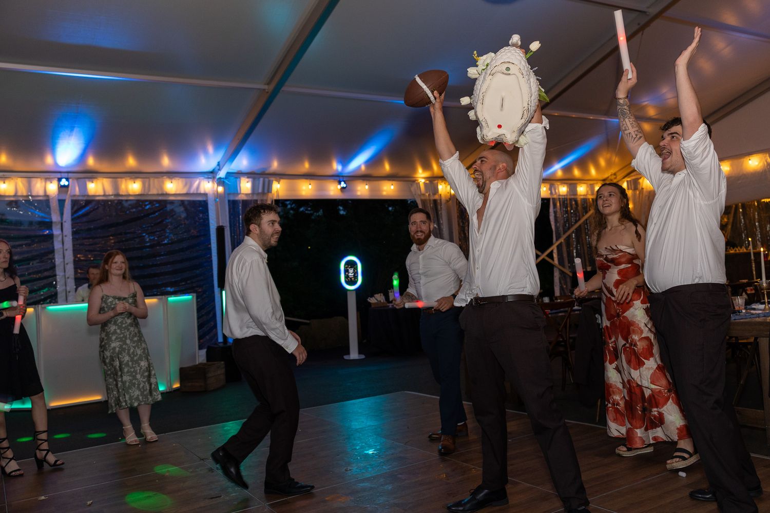 Men dancing and celebrating at a wedding reception under a tent with colorful lights.