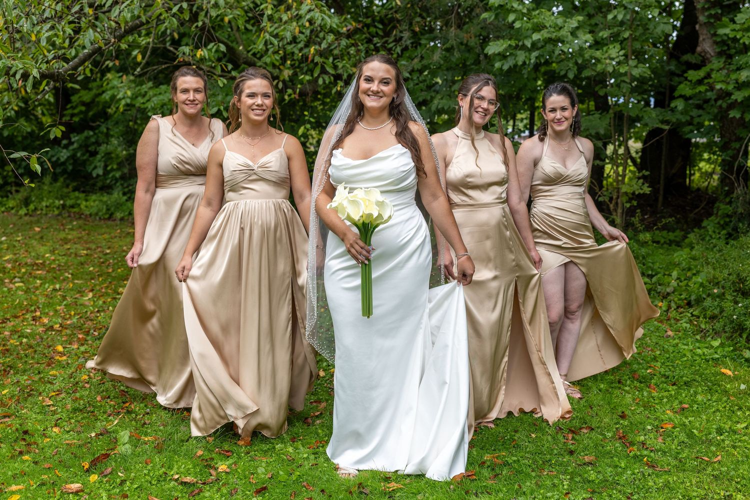 Bride in white gown holding bouquet poses with four bridesmaids in gold satin dresses outdoors on green lawn.