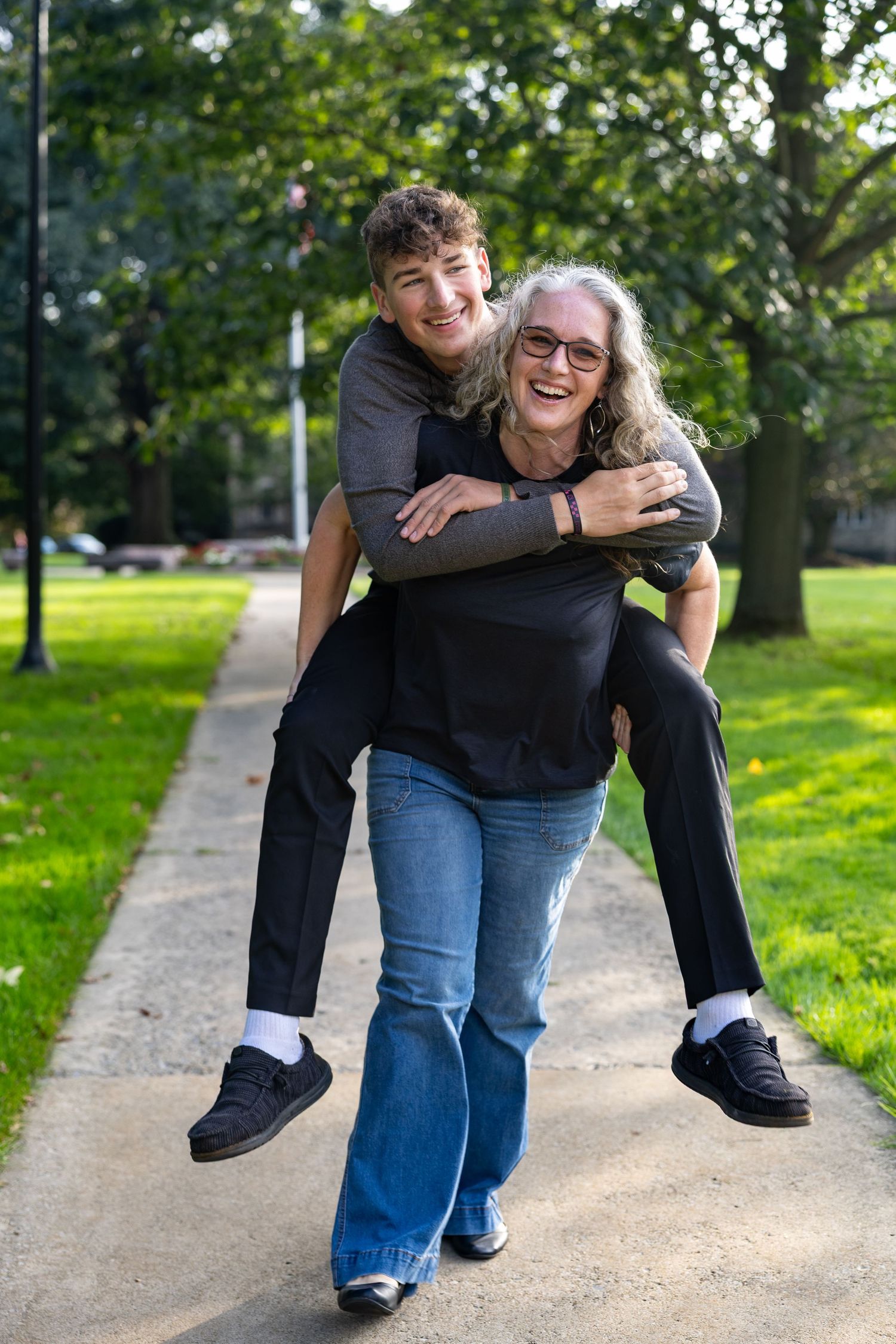 A woman gives a laughing teenage boy a piggyback ride on a park path surrounded by lush green trees.