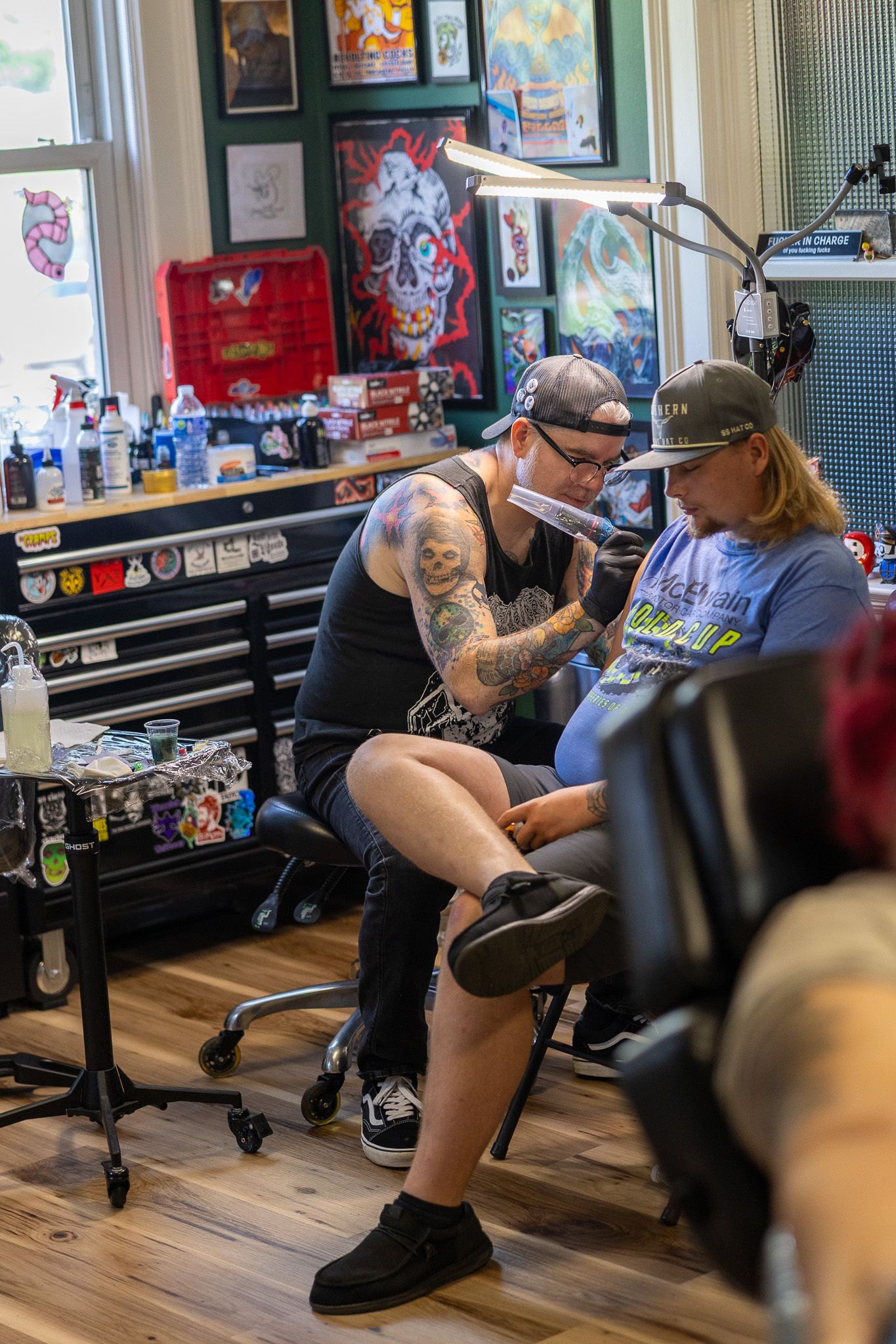 Heavily tattooed artist working on a clients leg tattoo under a bright lamp in a vibrant tattoo shop.