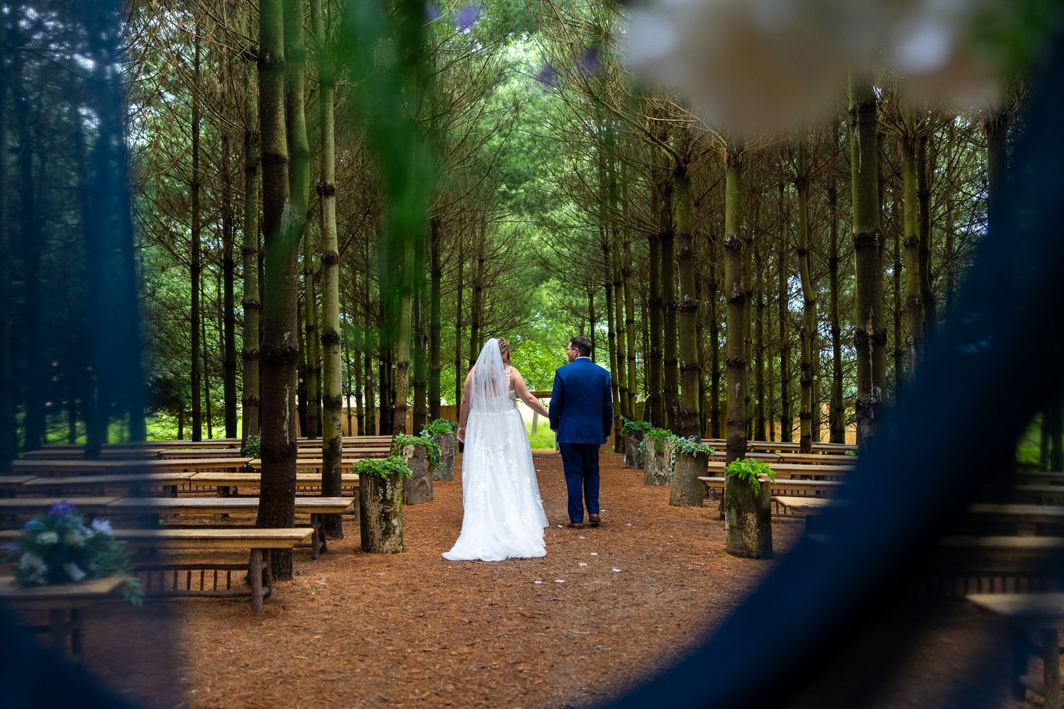 Bride and groom walk hand-in-hand down a tree-lined outdoor wedding aisle with wooden benches.