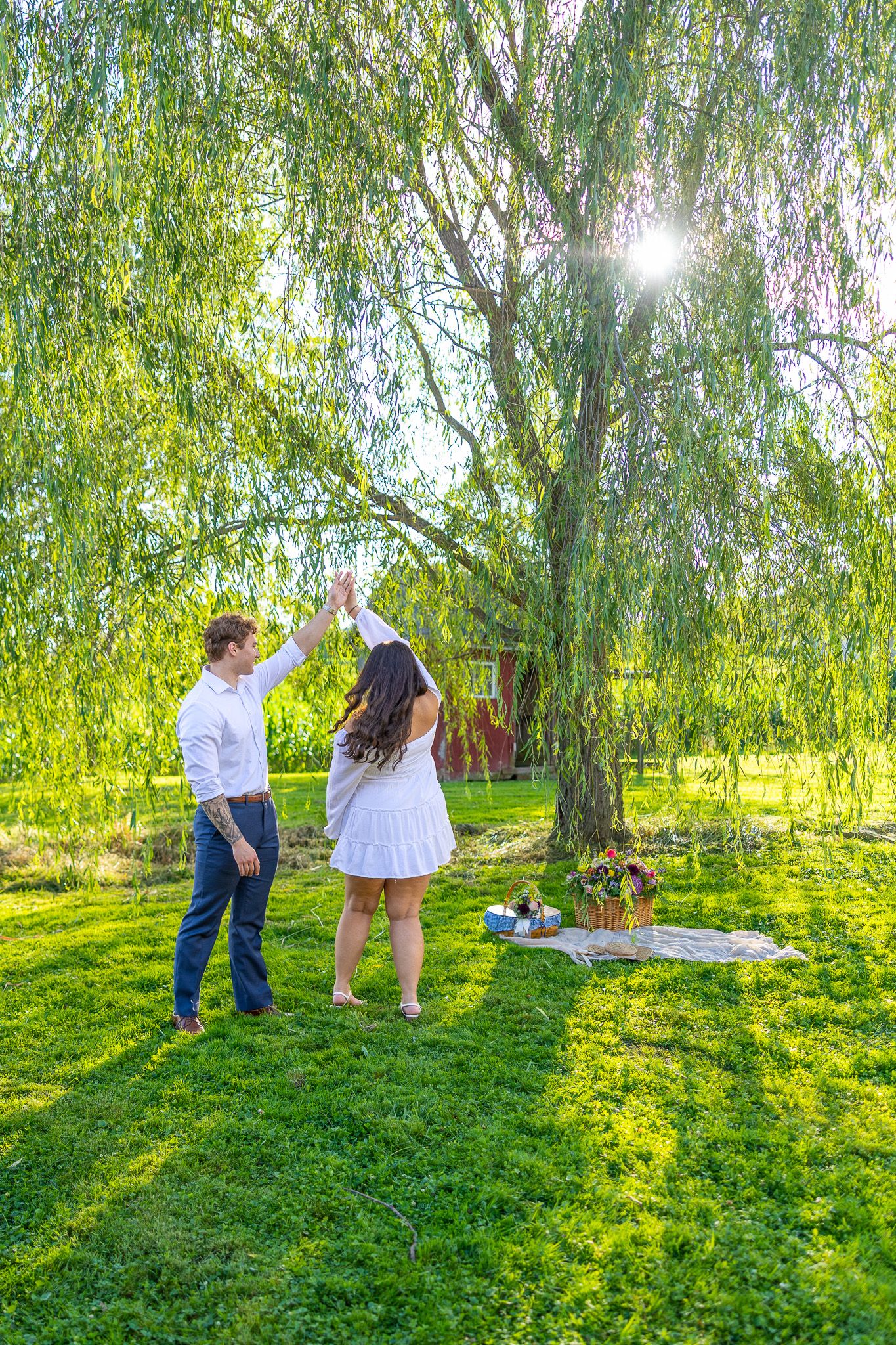 Couple dancing joyfully under a weeping willow tree in a sunny green park.