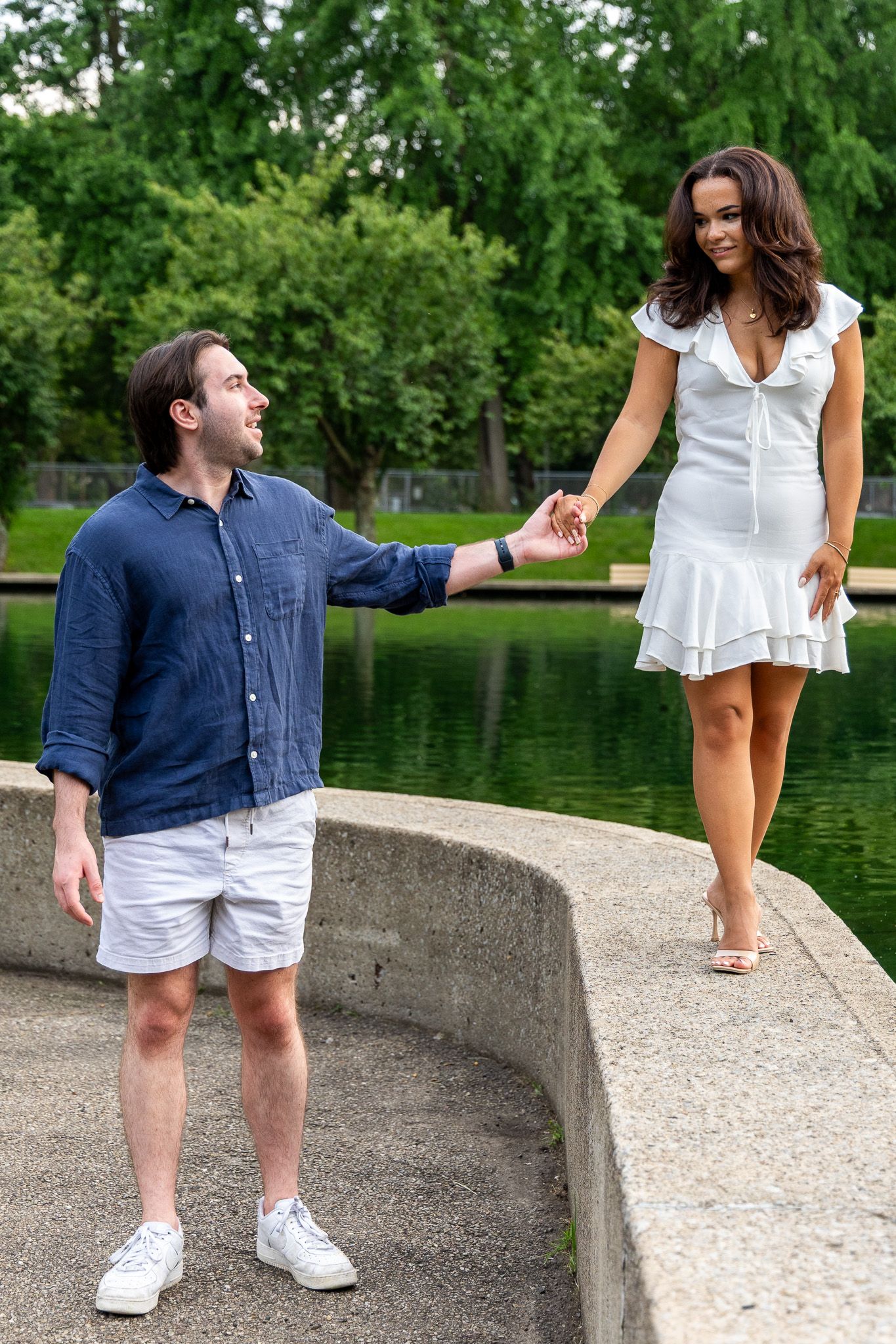 Couple holding hands near a reflective pond, woman in white dress leading man in blue shirt.